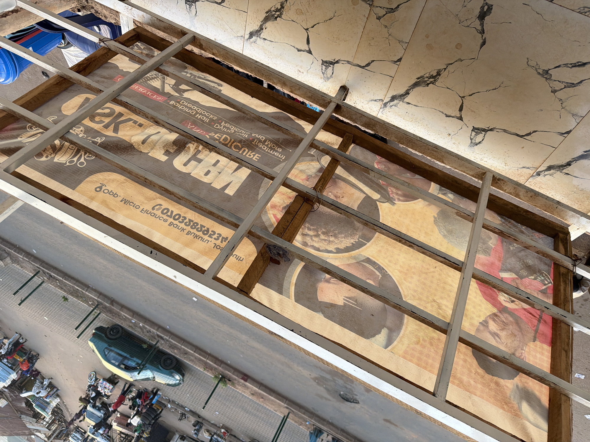This media file depicts an elevated, tilted view of an outdoor scene, likely in Bukuru, Nigeria. The primary focus is a large, transparent advertising banner stretched across a wooden and metal frame. The banner itself is printed with text and images, some of which are upside down relative to the viewer's perspective. Visible text includes "Disaqlocka," "STOM BRA," "ZEBAICER: Balpina Hall Dia," "VUR BEDICNBE," "СЕ СВИ," "Sobb WiCLO Luauce Bauk Boknin' סך," and a phone number "0103585023." The banner appears to feature stylized portraits or graphics.
Below and to the side of the banner, a street or pathway is visible, with a dark green vehicle overturned on its side in the middle of the road. The surrounding area shows signs of a marketplace or informal commercial activity, with various stalls or collections of goods and what appear to be people gathered around them. There are also some blue containers in the upper left portion of the frame, resting on a surface.
The lighting suggests it is daytime, and the weather appears to be clear. The cracked, tiled flooring in the upper portion of the image indicates a somewhat worn or aged environment. The overall impression is a snapshot of a busy, everyday scene in an urban or semi-urban setting, possibly capturing a moment of disruption due to the overturned vehicle.