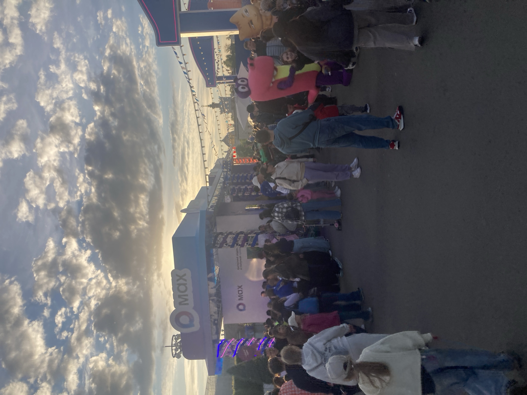 The image depicts a lively outdoor public event in Moscow, Russia, captured during the late afternoon or early evening with soft, golden light filtering through a cloudy sky. A large crowd of people, including adults and children, are visible, standing, walking, and possibly queuing within the event area.
Prominently featured are two costumed mascots among the attendees: one is a light brown/tan character with a rounded head, and the other is a distinctively large, pink, blocky figure with purple limbs and goggle-like eyes, seen interacting with a child. The crowd is dressed in casual clothing, including jeans, jackets, and hoodies.
The setting includes multiple temporary event structures or booths. A significant blue structure displays "Q MAX" (or "O MAX") in white lettering alongside a circular logo. A banner further back also features the "O MAX" logo. Partially visible Cyrillic text on one of the banners or screens reads "ВЫСТАВОЧНЫЙ ГОРОД КЛОУНСКОЙ", which translates to "Exhibition City Clown", suggesting a themed family entertainment or festival event. Bunting flags are strung overhead, enhancing the festive atmosphere. The ground appears to be paved, and in the distance, other buildings and trees indicate an urban or park environment. The scene conveys a sense of a busy, family-friendly public gathering.