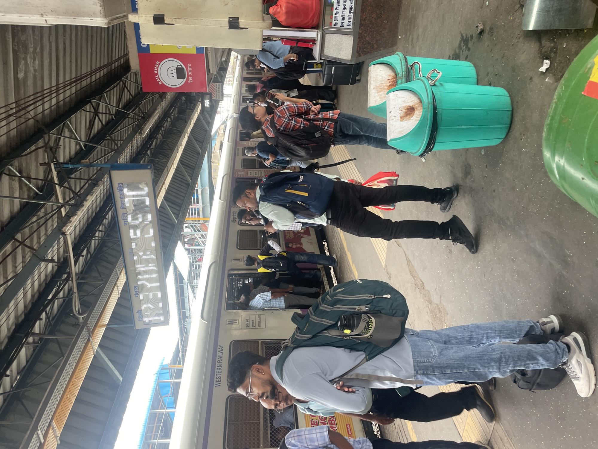 The image captures a bustling scene at a train station in Mumbai, India, specifically at the Western Railway platform. A train with "WESTERN RAILWAY" written on its side is stopped at the platform, with passengers disembarking and boarding. Several individuals are visible, many carrying backpacks and luggage, suggesting travel. The platform itself is paved and appears to be under a covered roof, characteristic of Indian railway stations. A digital display board showing train timings is visible hanging from the ceiling. Two green trash bins are prominently placed on the platform. The overall atmosphere suggests a busy and active public transport hub.