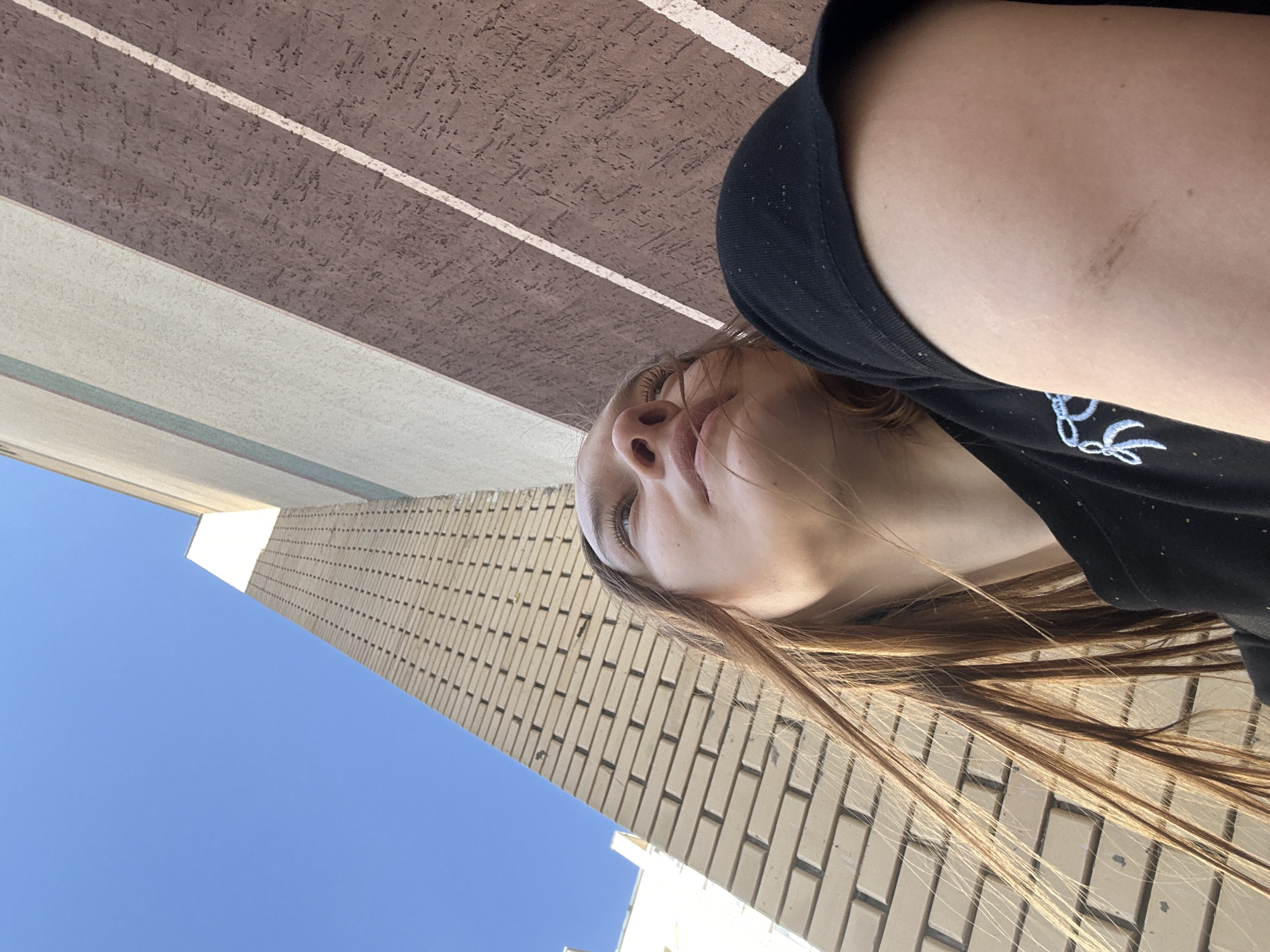 In an outdoor, low-angle, eye-level shot taken in Chaikovsky, Russia, a young woman with long, light brown hair looks up and to her right, her face angled towards the sun. She is wearing a black t-shirt with a light blue embroidered design. Her left arm is visible in the foreground, showing a small, dark mark on her skin. Behind her, a tall building with a brick facade stretches upwards, its light beige bricks contrasting with the bright blue sky. To the right of the brick building, another section of the building features a textured, reddish-brown facade with diagonal white stripes. The sunlight casts a warm glow on the scene.