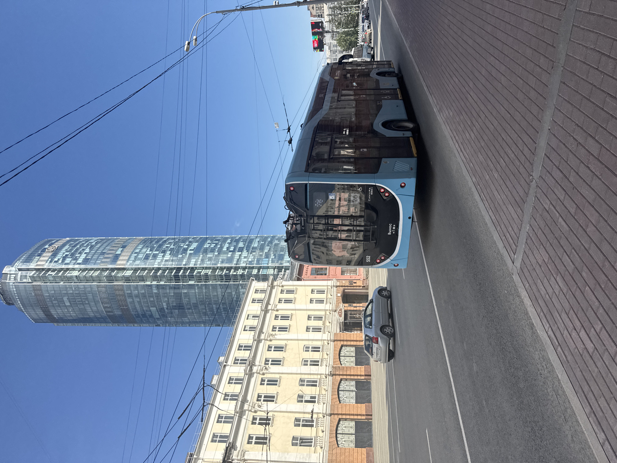 The image depicts a street scene in Yekaterinburg, Russia, on a clear day. A modern, light blue trolleybus with the number 552 visible on its rear is traveling on the right side of the road. The trolleybus is powered by overhead electric wires. To the left of the trolleybus, a silver car is also moving on the road. In the background, a tall, modern skyscraper with a glass facade dominates the skyline. Adjacent to the skyscraper and to the left of the road is a multi-story building with light-colored walls and numerous windows, exhibiting architectural features typical of older European city buildings. The street is paved with asphalt, and a brick-patterned sidewalk is visible on the right side. The sky is a clear, vibrant blue. The overall impression is that of a bustling urban environment with a mix of contemporary and historical architecture.