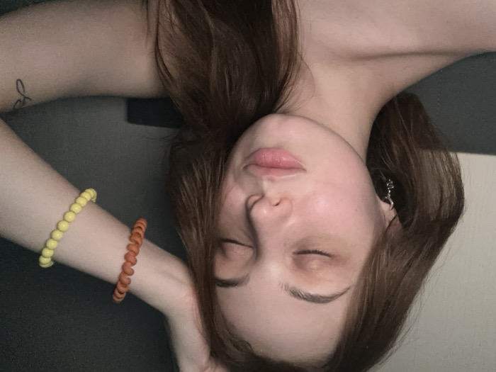 A close-up, chest-up image depicts a young adult female with long brown hair and fair skin. Her eyes are closed, and her lips are slightly parted. Her right arm is raised, with the hand positioned above her head, out of frame. She wears a silver-colored hoop earring with a small dark charm in her left ear. On her right wrist, two bracelets are visible: one orange, spiraled and textured, and one yellow, composed of uniform spherical beads. A small, black-ink tattoo resembling stylized script or a symbol is present on her upper left arm. The individual appears bare-shouldered. The background is dark and indistinct, indicative of an indoor setting with subdued lighting. There are no visual identifiers within the image to confirm a specific location in Kommunarka, Russia.