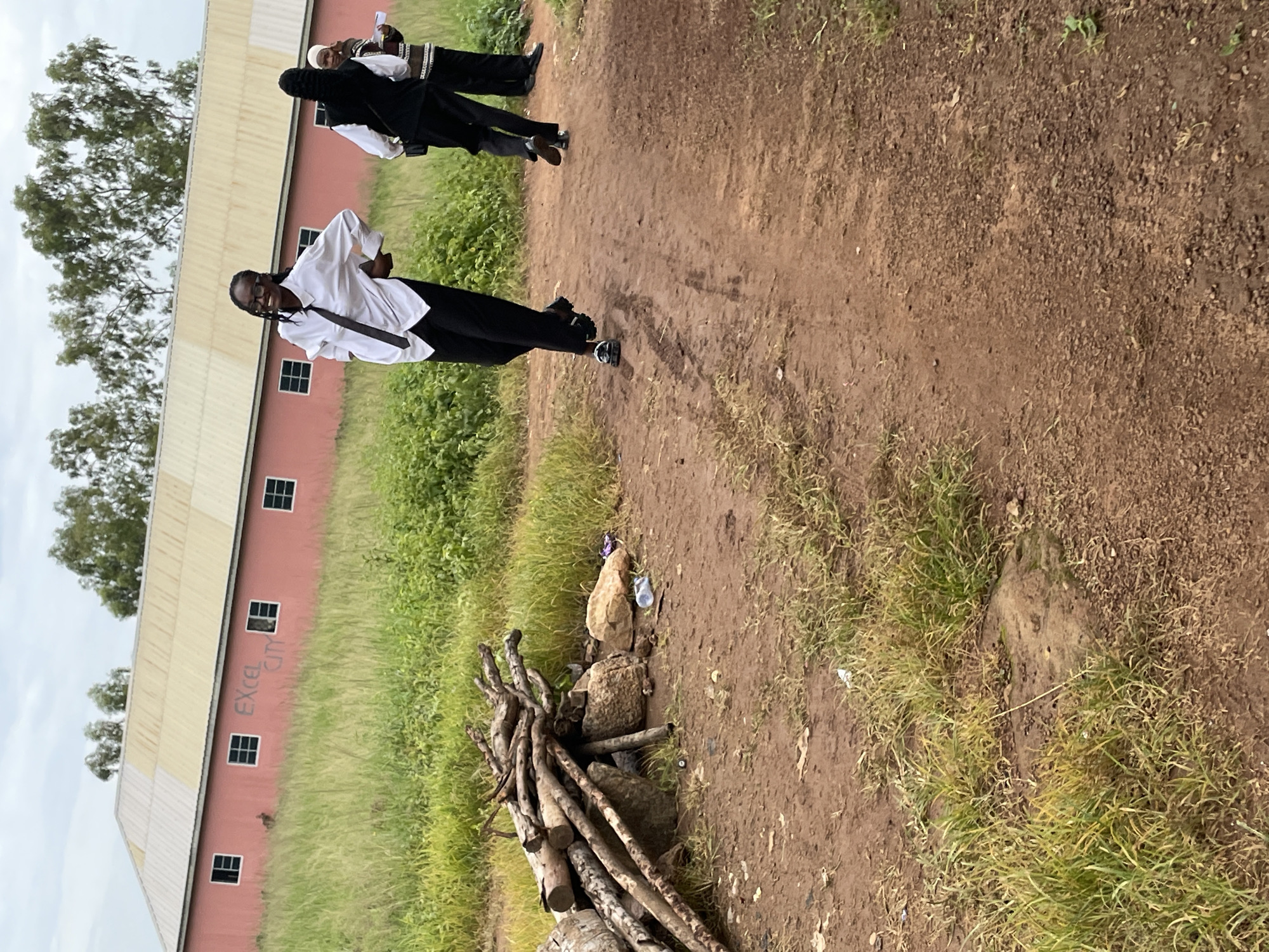 In Durumi, Nigeria, a group of individuals, appearing to be students in formal attire, are captured outdoors. The scene features a large, pale yellow and pink building with multiple windows, suggestive of an educational institution. The building has the words "EXCEL CITY" visible on its side. In the foreground, a pile of logs rests on the ground, accompanied by scattered rocks and a plastic bottle, indicating a somewhat unkempt environment. The ground is a reddish-brown dirt path with patches of green grass. The individuals are walking on this path. One person in the center, wearing a white shirt, black tie, and black pants, is looking towards the camera with a smile and has their hands on their hips. Two other individuals in similar attire are walking away from the camera. The overall atmosphere suggests a moment captured during or after an event at the school.
