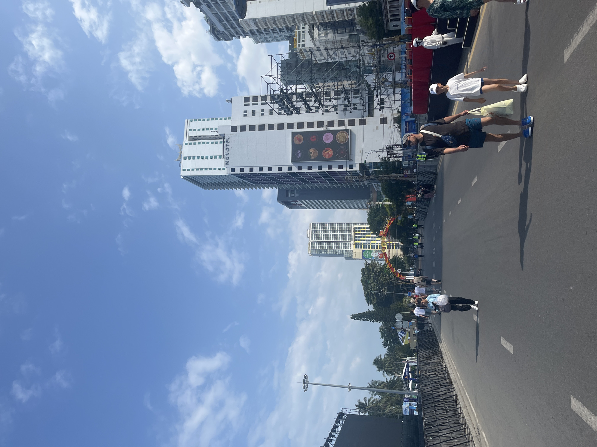 The image depicts a street scene in Nha Trang, Vietnam, likely during an event or festival. The sky is blue with scattered clouds. Tall buildings, including a Novotel hotel, dominate the background, indicating an urban environment. In the foreground, a paved road with white lane markings is visible. Several people are walking on the road. A man and a young boy, both wearing casual attire, walk towards the viewer, with the boy carrying a plastic bag. Further down the road, a woman in black pants and a light-colored top is walking away from the viewer. On the left side of the road, a large black screen or stage structure is set up, along with a black metal fence. On the right side, there's a red barrier and scaffolding, suggesting construction or event setup. An archway decorated with colorful banners spans across the road in the mid-ground. Palm trees are visible among the buildings and foliage, characteristic of a coastal city. The overall impression is a lively, sunny day in a bustling Vietnamese city.