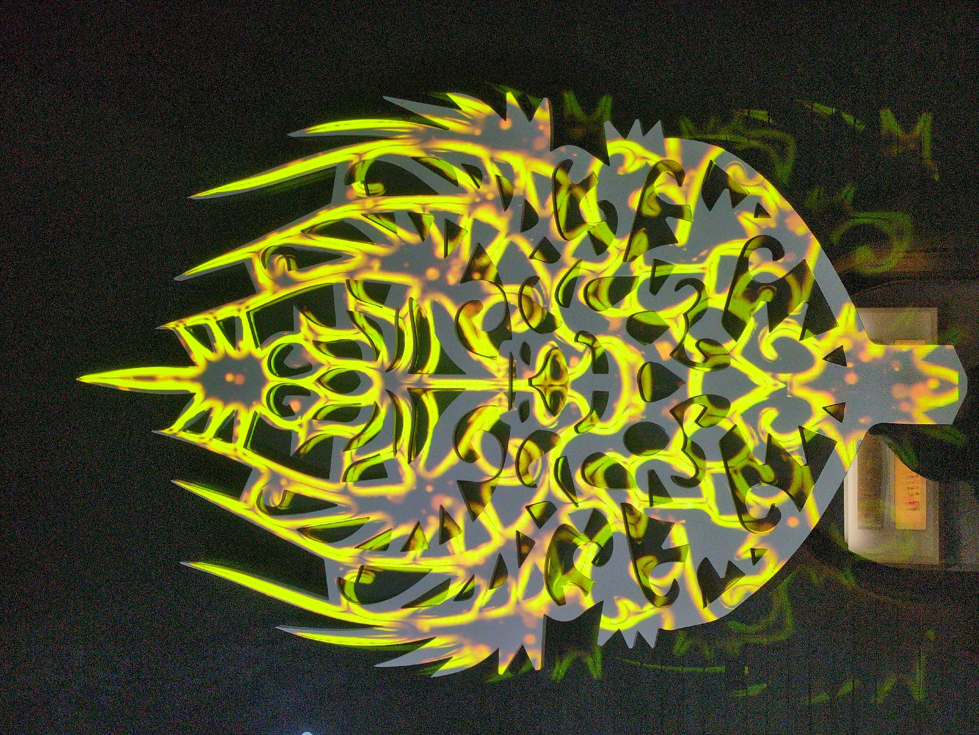 A close-up shot shows a decorative cutout of a stylized fish shape, illuminated with bright, yellow, abstract patterns that resemble glowing symbols. The cutout is light gray and has intricate designs within its boundaries. The background is dark, grainy, and out of focus, suggesting a low-light environment. A faint, greenish light is visible in the upper right quadrant of the image, and the scene appears to be indoors. Details indicating the specific location within Gongju-si, South Korea, are not evident.