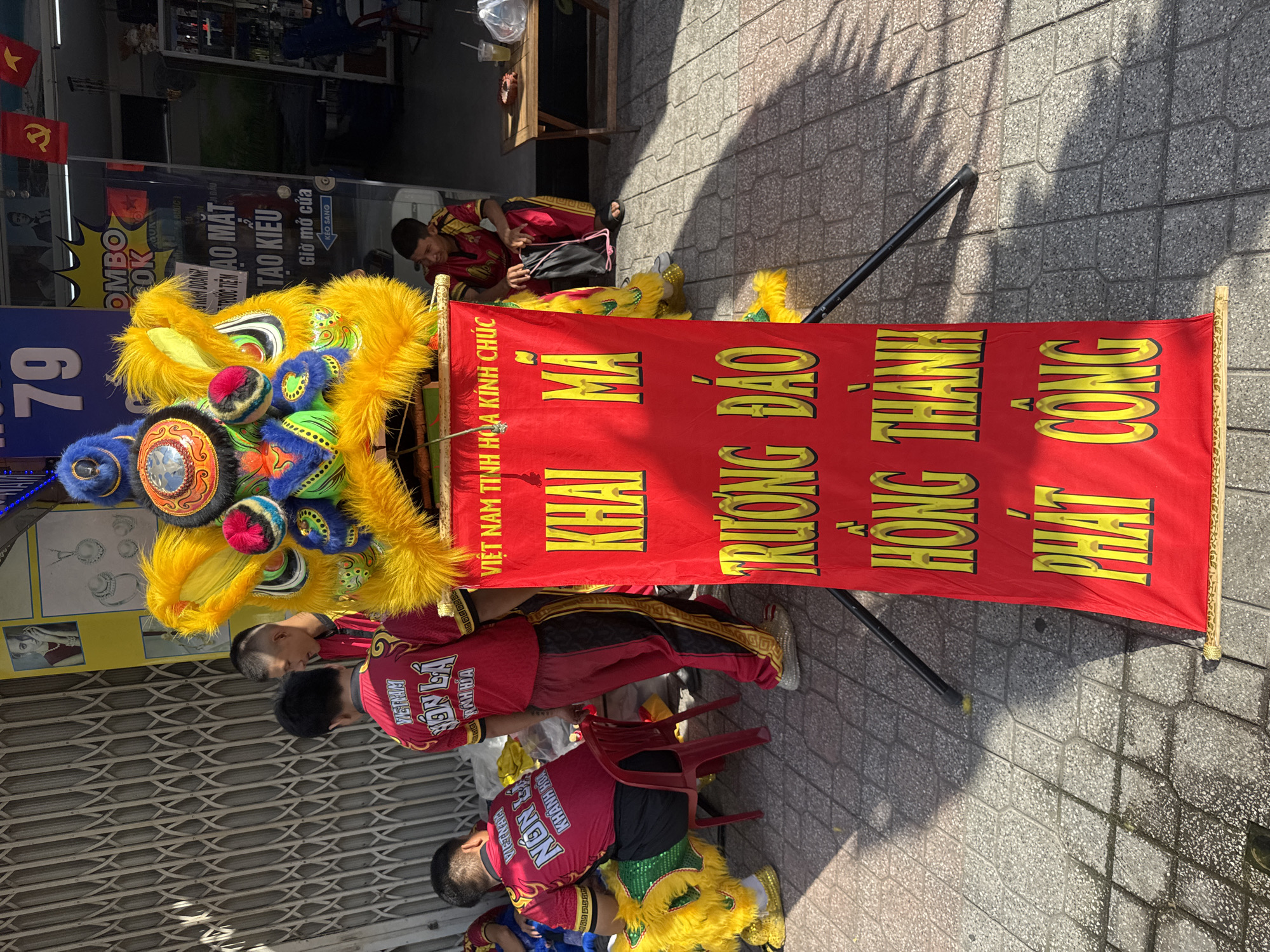 This is a medium shot of a street scene in Nha Trang Ward, Vietnam, featuring a vibrant red banner with yellow lettering, propped up by two black stands. The banner's text, in Vietnamese, reads: "VIET NAM TINH HOA KINH CHUC KHAI MA TRUONG DAO HONG THANH PHAT CONG". In front of the banner is a colorful, fluffy lion dance costume, predominantly yellow with blue, green, and red accents. To the left of the banner, two individuals are visible. One, wearing a red shirt with "VIET NAM NON LA" and "KHANH HOA" printed in white, is seated on a red plastic chair. Another person in a similar red outfit, with black pants featuring a gold pattern, stands behind him. Further to the right, a man in a red and gold outfit is seated near a small wooden table with a drink and a plastic bag on it. The background shows a building with a closed metallic gate on the left and shop windows on the right, displaying various items and posters, including a number "79" prominently featured on one of the shop fronts. The ground is paved with textured tiles, and the scene is bathed in sunlight, casting shadows.
