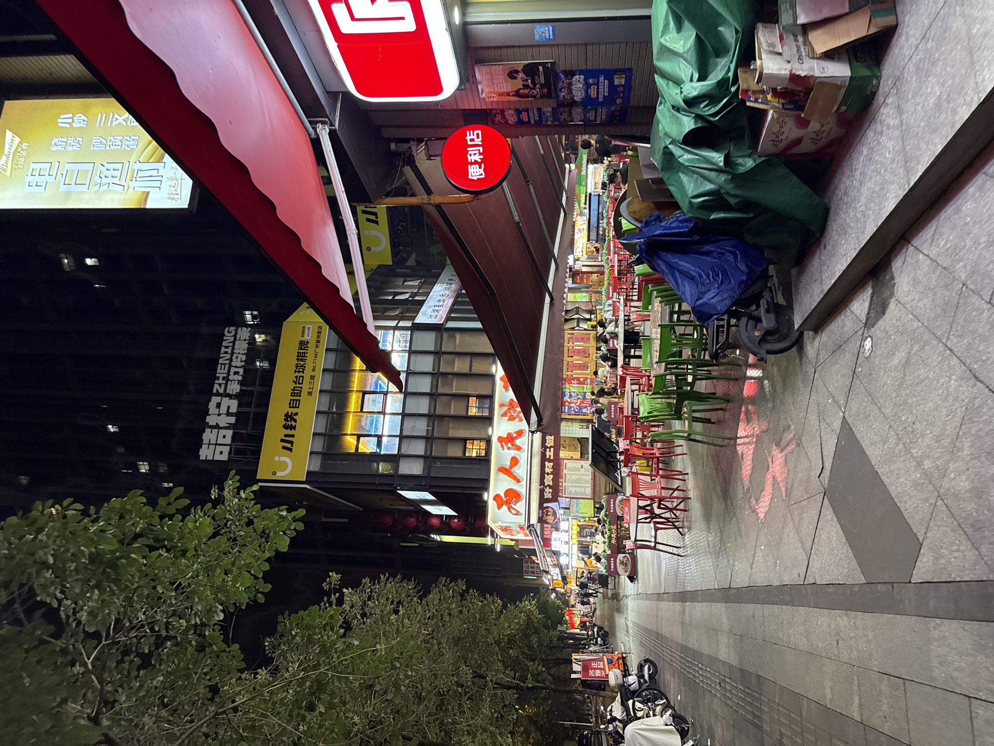 This is a nighttime outdoor shot of a bustling street scene in Haizhu District, China. The image is vertically oriented. On the left, there's a building with several floors, its windows dimly lit. A red awning extends from the building, casting a shadow. Below the awning, a sign with Chinese characters and the Budweiser logo is visible.
The street itself is lined with restaurants and shops, indicated by numerous brightly lit signs in Chinese. Many of these signs advertise food and drinks, with text such as "烧烤 砂锅粥" (barbecue, claypot porridge) and "手打柠檬茶" (hand-beaten lemon tea). Red lanterns hang from some establishments, adding to the festive atmosphere.
In the foreground and extending into the middle ground, several tables and chairs are set up outside, suggesting an outdoor dining area. The chairs are predominantly green and red. In the background, more shops and diners can be seen, with people sitting at tables. A blue tarp is covering some items, and a green tarp is draped over a pile of boxes and other objects on the right side of the street.
On the right side of the street, a paved walkway is visible, with a dark line running along its length. Several motorcycles are parked on the sidewalk, some covered with white sheets. The overall impression is one of a lively commercial street at night, likely a popular spot for locals to eat and socialize. The presence of neon signs and the general ambiance suggest it's a warm evening.