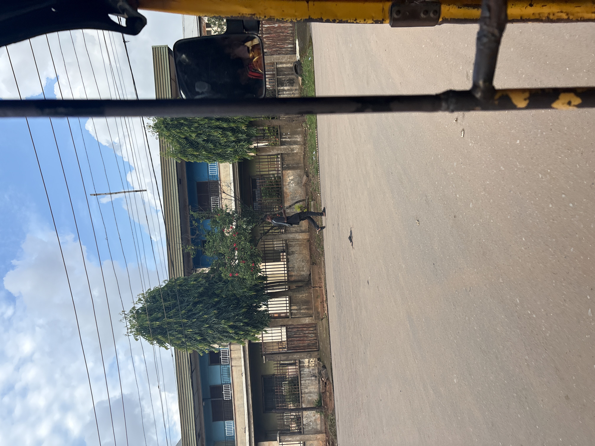 This image depicts a street scene in Onitsha, Nigeria. The foreground is dominated by the yellow and black bars of what appears to be a vehicle, possibly an auto-rickshaw, with a side mirror reflecting a blurry image of a person inside. The middle ground shows a paved road with a single person walking from left to right, wearing dark clothing. Behind the road is a fenced property with a two-story building. The building has a light blue upper level with balconies and a darker lower level. Several trees, including tall, slender evergreen trees and a tree with red flowers, are visible within the property. The background shows a cloudy sky with power lines crisscrossing overhead. The overall environment suggests an urban setting with residential or commercial buildings lining the street.