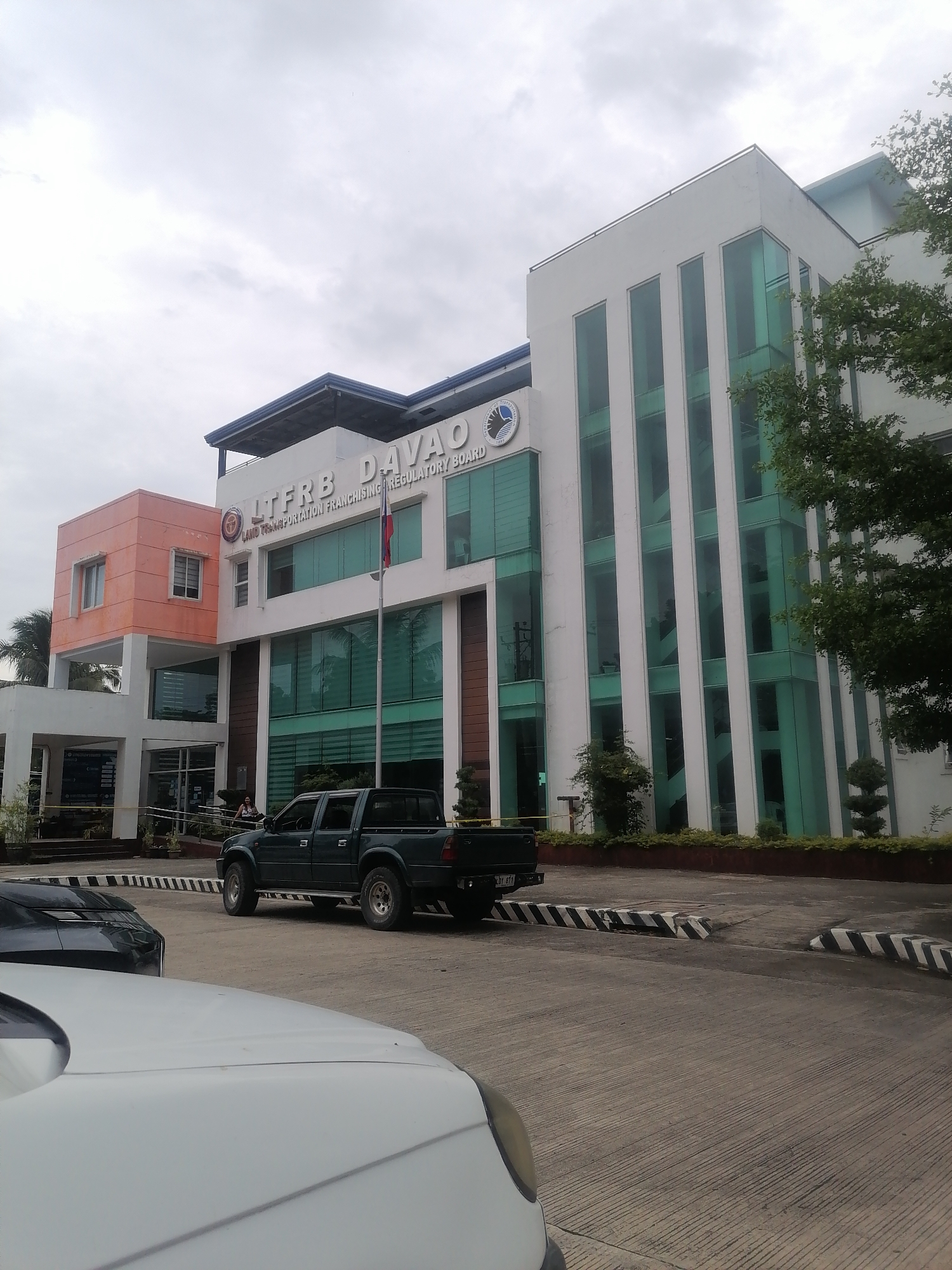 This image captures the exterior of a modern multi-story building, identified by its signage as "LTFRB DAVAO" and "LAND TRANSPORTATION FRANCHISING REGULATORY BOARD," confirming its location in Davao City, Philippines. The building features a prominent white section with large, vertically-oriented green-tinted glass windows, and an adjacent orange-colored section with smaller, rectangular windows. A Philippine flag flies from a flagpole in front of the main entrance.
In the foreground and midground, several vehicles are visible. A dark green pickup truck is parked or moving on the concrete driveway. Parts of two other cars, one dark and one white, are partially visible in the immediate foreground, suggesting a parking area or drop-off zone. The ground features concrete with etched lines and black-and-white striped speed bumps or curb markings.
The sky is overcast, indicating a cloudy daytime setting. Lush green foliage from trees and bushes flanks the building and driveway. One person can be faintly seen near the building's entrance. The scene depicts a typical administrative office building environment, possibly during business hours.