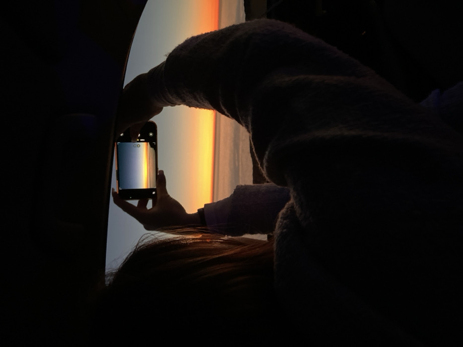 The image depicts an individual from behind, seated inside a vehicle, silhouetted against a multi-hued sunset or sunrise over a body of water. The person, with visible light-brown hair and wearing a light-colored, textured long-sleeved garment, holds a black smartphone horizontally with both hands, positioned to photograph the external scene. The smartphone's screen displays a sky gradient from dark blue to yellow and orange, meeting a hazy horizon above the water. The upper frame of a vehicle window is visible, with the interior predominantly in shadow due to the strong backlighting from the low sun. The scene depicts a coastal environment, viewed from an elevated position overlooking the ocean or sea at either dawn or dusk. The location in the city is Unknown, Unknown.