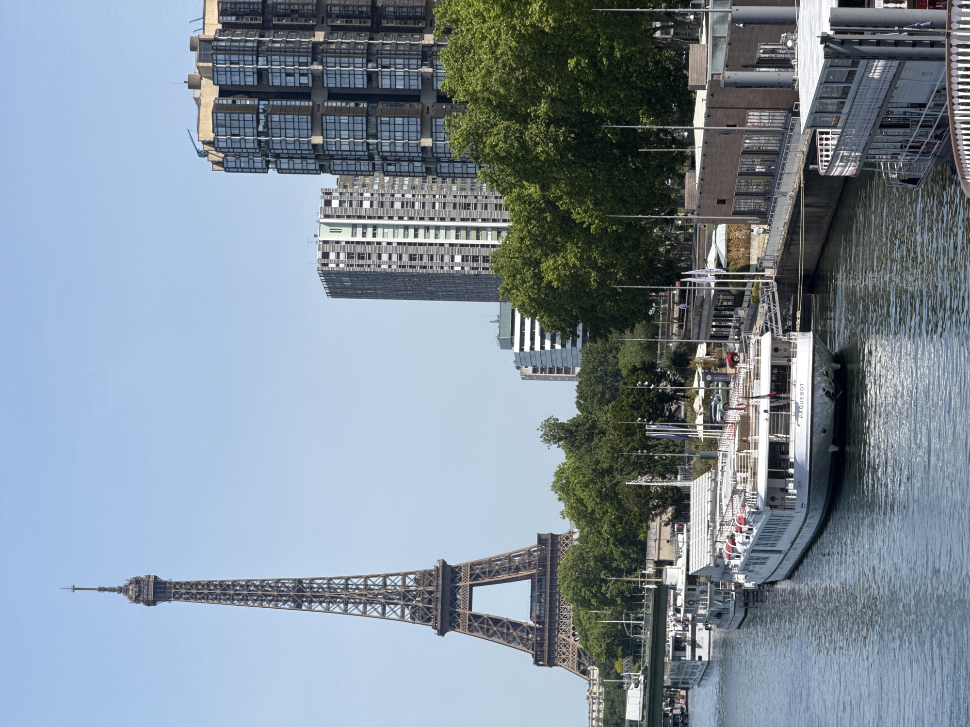 A long, vertical outdoor shot shows the Eiffel Tower on the left, partially obscured by trees along the center of the shot, alongside a river with tourist boats docked. Above the trees are various apartment buildings and office towers. The weather is clear and sunny. The Eiffel Tower is a detailed, wrought-iron lattice structure and is brown. The trees have dark-green foliage. The tour boats are white with passengers boarding. The river water reflects the sky and trees. The buildings are a mix of light-gray, brown, and glass. The buildings are mostly box-shaped and are of medium height, except for the Eiffel Tower.
