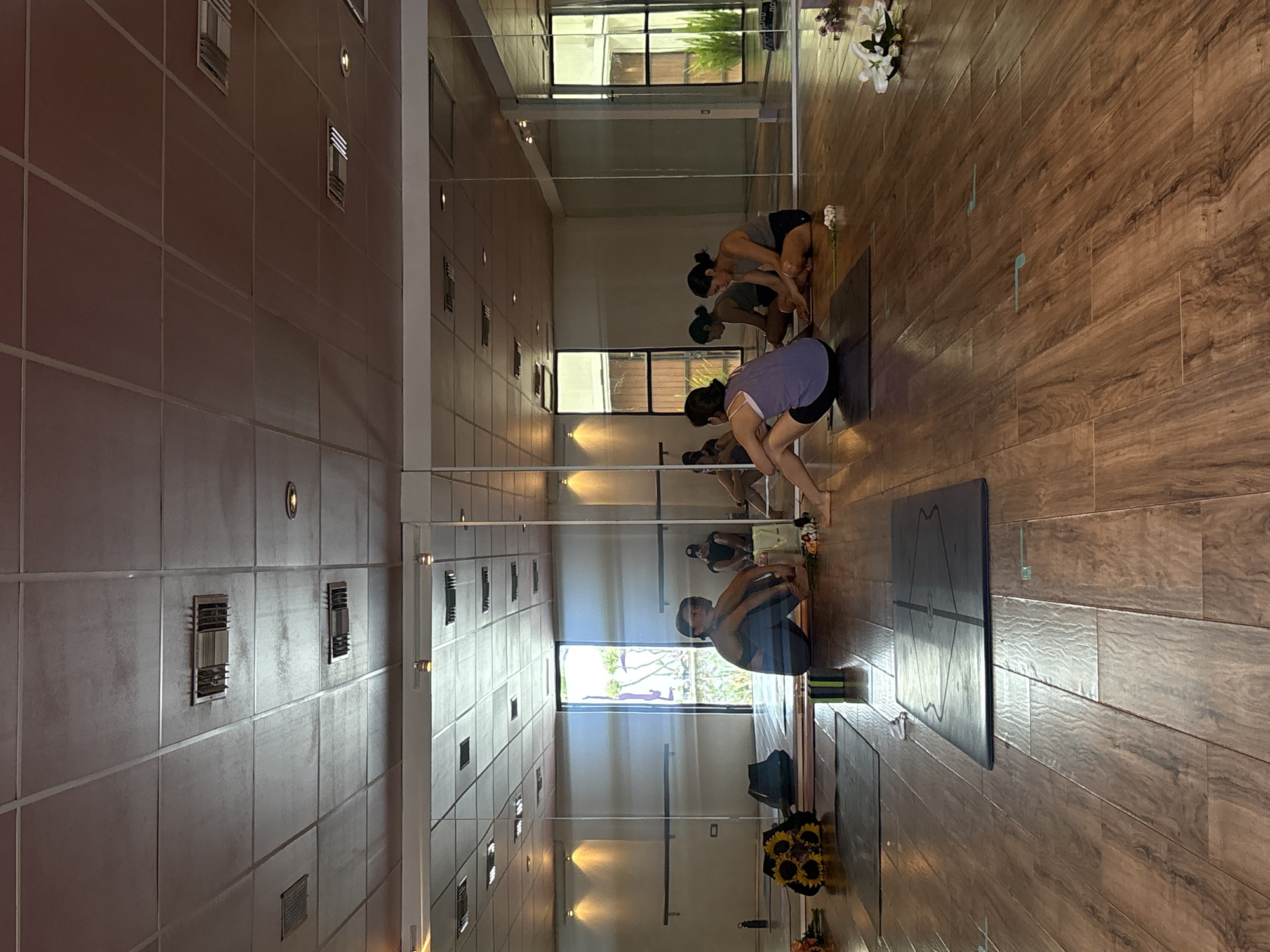 The image captures a yoga studio setting, likely in the city of Puebla, Mexico. The studio is lined with floor-to-ceiling mirrors, creating multiple reflections of the scene.
There are several people in the studio, most of whom are engaged in yoga poses on mats. The individuals are positioned facing forward in the room, bent over in what appears to be a forward fold.
The floor is covered with wood-grain tiles. There are yoga mats placed on the floor in front of each person, and some mats have geometric designs. A bag and a bouquet of flowers are placed in the bottom corner.
The ceiling of the studio features a grid-like pattern. The walls appear to be tiled, and there are ventilation grates. The room is well-lit, primarily from natural light entering through a window at the far end. The window provides a view to greenery outside, likely trees or plants.