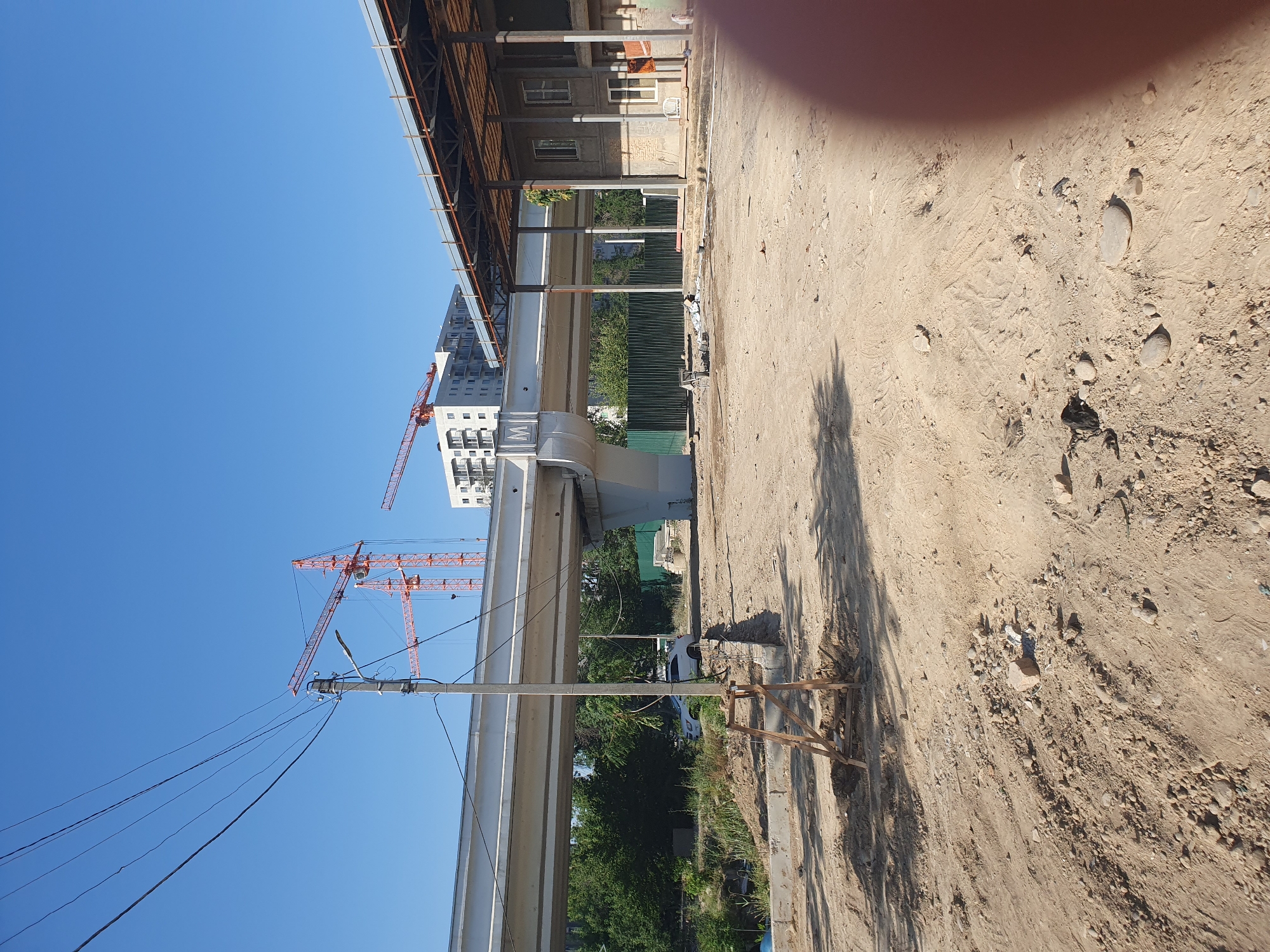 The image depicts a construction site in Tashkent, Uzbekistan, under a clear blue sky. In the foreground, a dirt and gravel area shows evidence of recent work, with tire tracks visible in the sand. A makeshift wooden support structure is partially buried in the ground.
Dominating the mid-ground is an elevated concrete structure, likely part of a transportation or infrastructure project. The letter "M" is prominently displayed on one of its pillars. To the left of this structure, a utility pole with power lines stretches across the sky, and a streetlamp is attached to it. Behind the concrete structure, trees and foliage are visible.
In the background, several orange construction cranes are operating, suggesting ongoing development. A modern, multi-story building stands to the left, and another partially constructed building with a covered roof and support columns is on the right. A white car is parked near the elevated structure. The overall scene suggests a large-scale urban development project in progress.