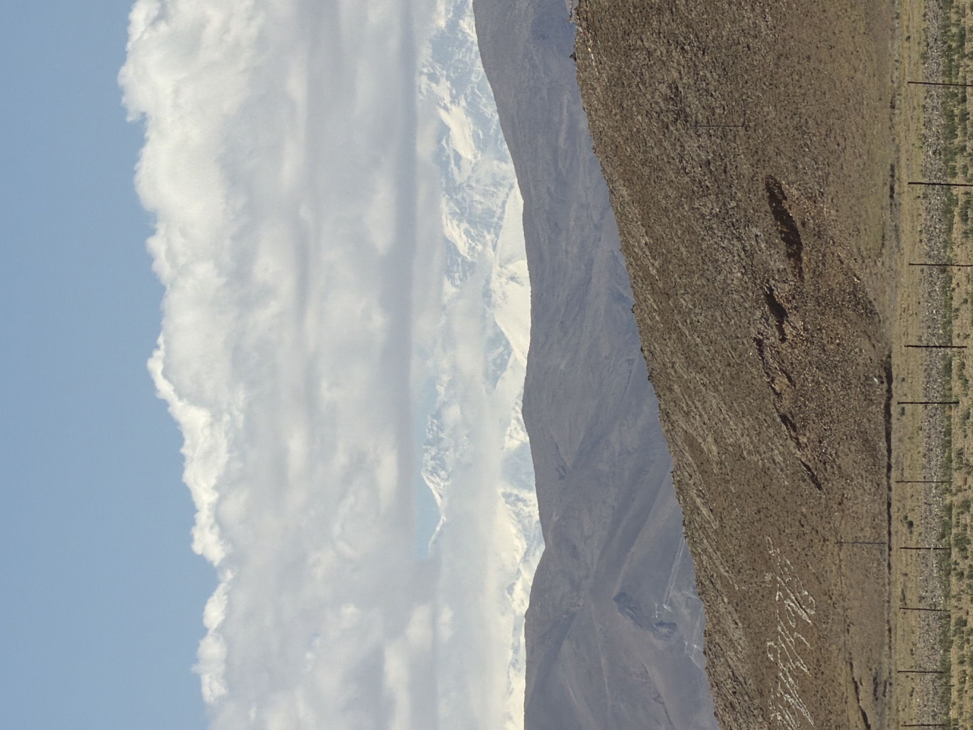 A landscape photograph captures a vast, mountainous, and arid region under bright daylight. The image is dominated by layers of geological formations and sky.
On the far right, a steep, barren hillside slopes upwards, covered in brown and reddish-brown rocky terrain with sparse, dry vegetation. Along the bottom edge of this slope, a narrow, unpaved track or path runs horizontally, lined with several dark, thin, regularly spaced vertical posts or markers. On the upper part of this foreground hillside, large white letters are inscribed or laid out, clearly spelling out "XINJIANG" in a stylized, somewhat cursive font.
Immediately to the left of the foreground hillside, a range of darker, smoother, and more muted purplish-grey mountains extends across the mid-distance. Further in the background, to the left of these mountains, majestic snow-capped peaks rise, partially obscured by a thick bank of white cumulus clouds that stretch vertically across the central-left portion of the frame.
The far left of the image features a clear, bright blue sky, gradually giving way to the fluffy white clouds that envelop the distant mountain tops. The sunlight is strong, indicating a clear day, and the overall scene suggests a high-altitude, potentially desert-like environment. No people or specific activities are visible, but the inscribed text points to human presence and marking of the landscape.