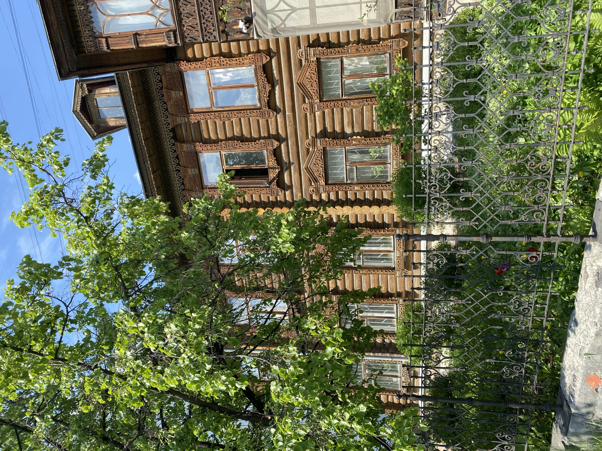 The image captures a wooden building in Yekaterinburg, Russia, partially obscured by a tree and a wrought-iron fence. The building's facade features traditional Russian wood carving around the windows and along the edges. The tree, with its lush green leaves, is positioned to the left of the building. The wrought-iron fence, with its ornamental patterns, is in the foreground, partially blocking the view of the garden behind it. The sky, a clear blue, is visible in the upper left corner.