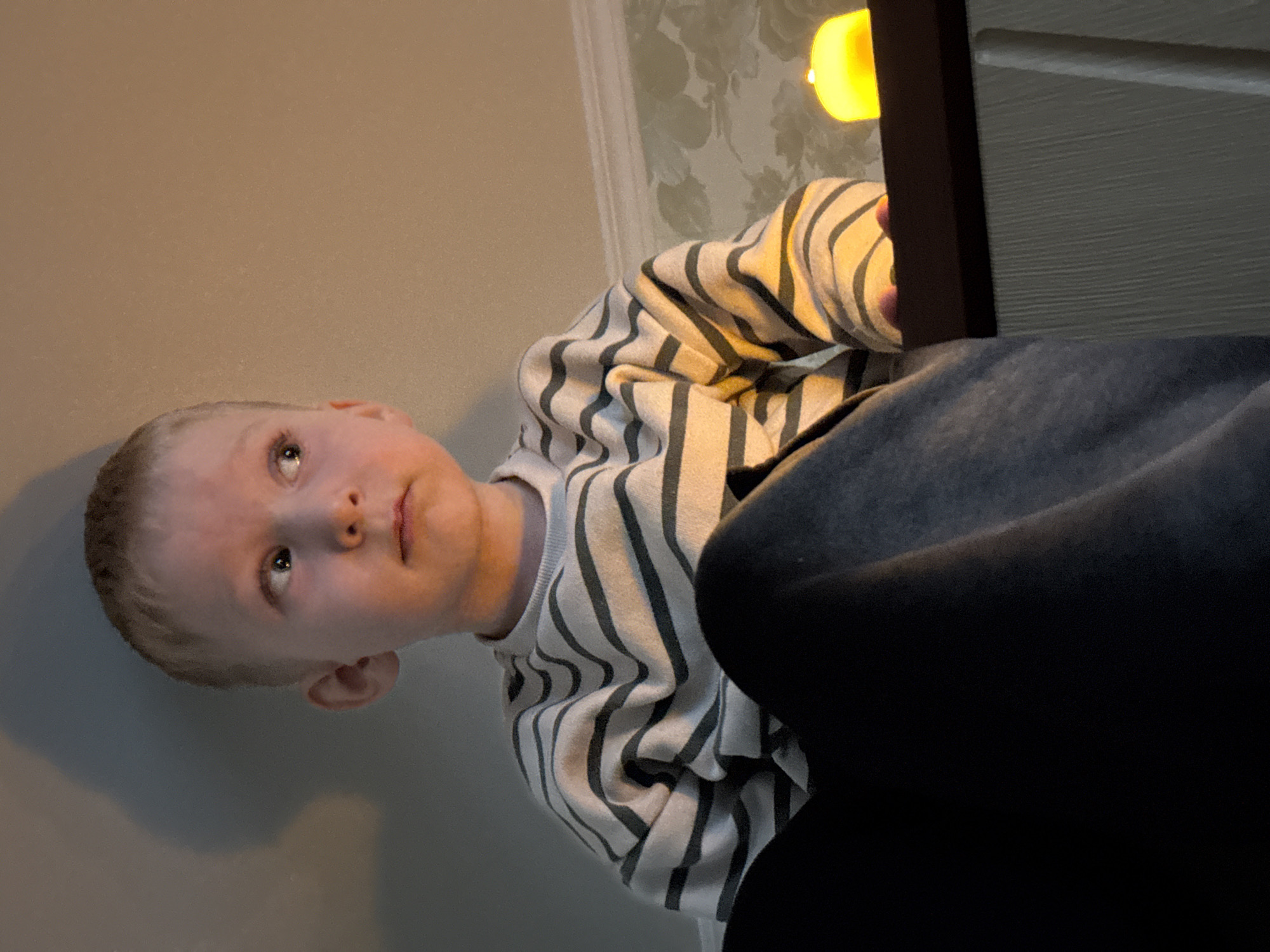 A young, fair-skinned child, likely a boy with short, light hair, is captured in a close-up, upward-looking shot. The child's eyes are light-colored (possibly blue or grey) and are directed thoughtfully upwards and slightly to the right, suggesting engagement with something above their line of sight. They wear a light-colored long-sleeved sweater adorned with dark grey or black horizontal stripes.
The child appears to be holding a dark, rectangular object, possibly a tablet or a book, which emits a bright yellow light. This light source illuminates the child's fingers and casts a warm glow, suggesting the object is actively displaying content or is a lamp. The rest of the child's body is partially visible, clothed in dark trousers or leggings, which are prominent in the foreground and lower right of the image.
The setting appears to be indoors. Behind the child, a wall covered with subtle, light-colored floral or leafy patterned wallpaper is visible on the right, while the left side of the background shows a plain, neutral-colored wall (beige/light brown). The overall lighting suggests an indoor environment, possibly a home, where the artificial light from the object is a dominant illumination source. There is no visible text in the image. The child's expression is one of focused curiosity or contemplation. The external context places the location in Perm, Russia.