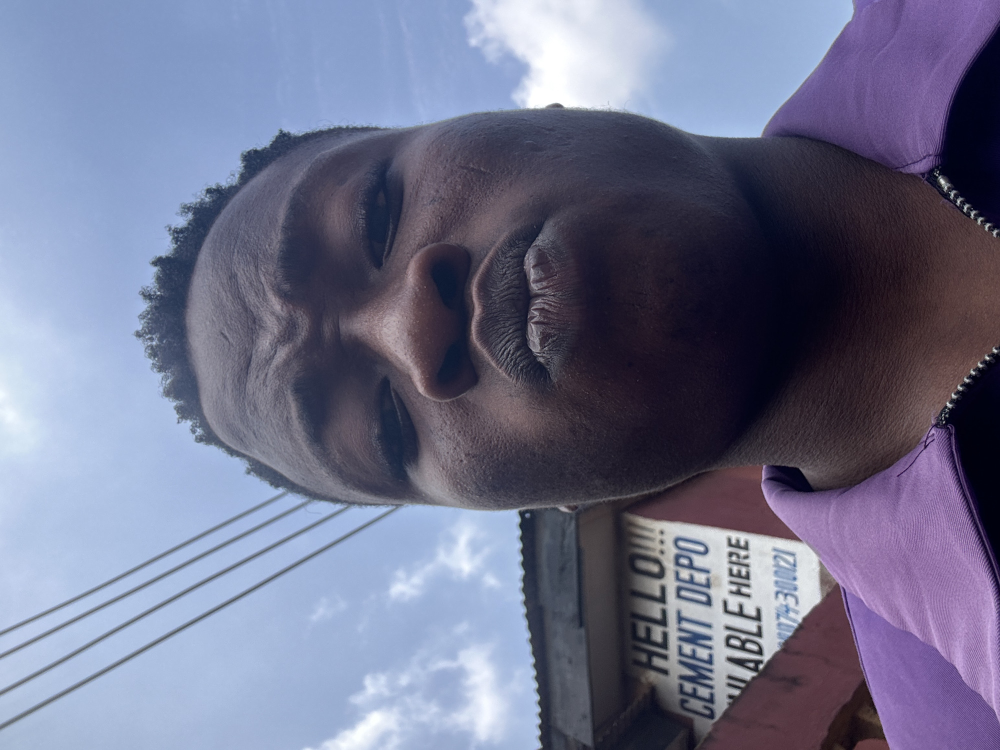 The photograph shows a close-up selfie of a young man with dark skin and short, curly hair. He is wearing a purple jacket with a silver zipper. The angle is from below, looking up at his face, which is close to the camera. His expression appears to be frowning or serious.
Behind him, partially out of focus, is a sign reading "HELLO!!! CEMENT DEPO AVAILABLE HERE" and a phone number "079-300021". The sign appears to be on the side of a building with a corrugated metal roof. Power lines can be seen against a blue sky with scattered clouds.
Based on the cement depot sign, the location is likely in the city of Ikorodu, Nigeria.