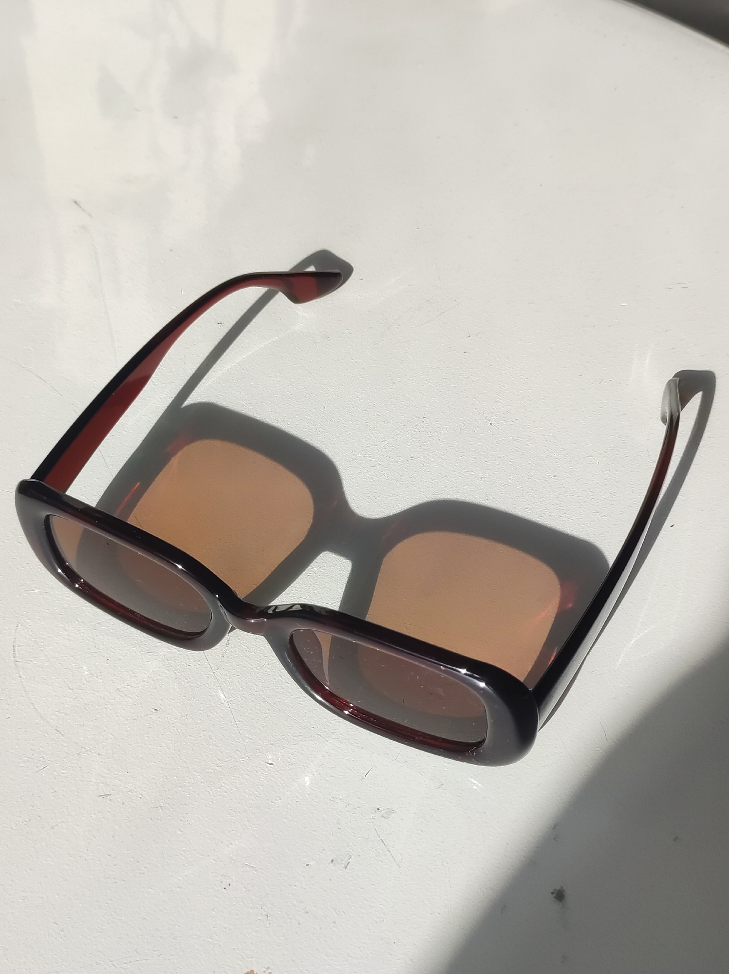The image shows a pair of rectangular sunglasses with a dark brown frame and light brown lenses lying on a white, possibly outdoor surface. The sunglasses are casting a shadow that mimics their shape onto the surface. There is no information present to suggest that the location is Chaikovsky, Russia.