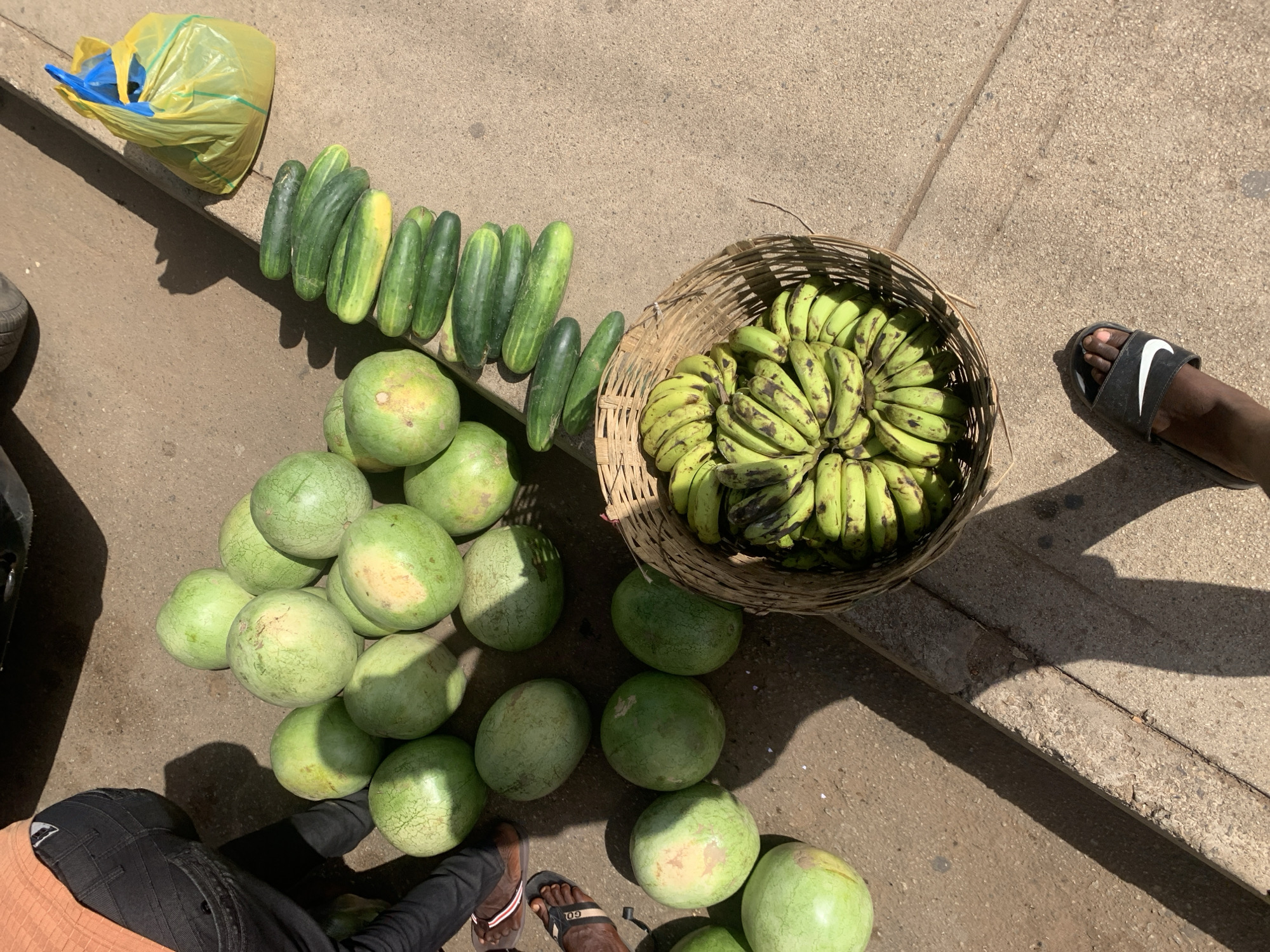 The image shows a street-side fruit and vegetable vendor in Jos, Nigeria. Arranged on the pavement are a variety of items for sale: a cluster of green watermelons, a row of cucumbers, and a woven basket filled with green bananas. A yellow plastic bag sits next to the cucumbers. A person wearing black trousers and brown sandals is seen near the watermelons. Another person wearing a black slide sandal with a white Nike logo is standing close to the basket of bananas. The pavement is cracked and discolored in places.