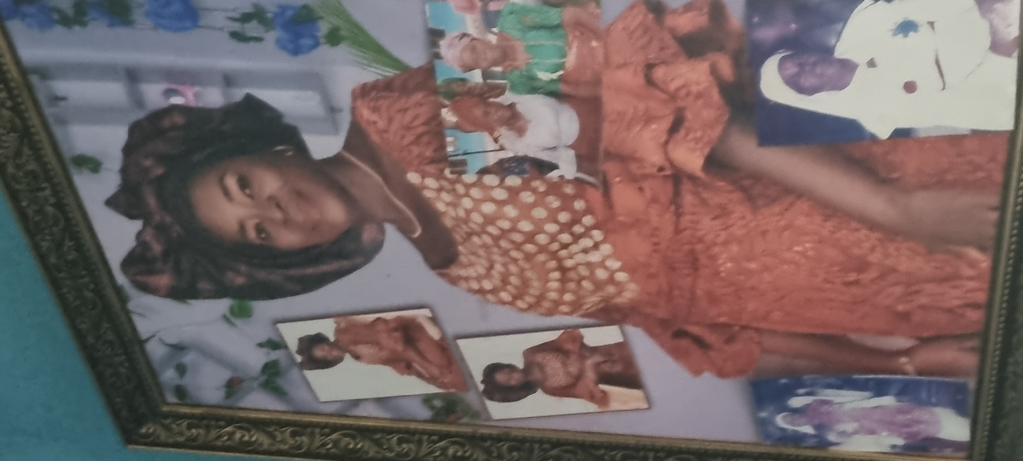 The image is a framed photograph, likely an 8x10 or larger, with a distressed gold-colored ornate frame. The photograph itself depicts a collage of multiple images, with a central, dominant portrait of a Black woman. She is smiling slightly and wearing a large, elaborate headwrap and an orange lace dress with a white polka-dot overlay on the bodice. There are several smaller inset photographs within the larger frame.
To the left of the central portrait are two smaller, vertical rectangular photos, also of the same woman in similar attire. Below and to the right of the central portrait are two more smaller, horizontal rectangular photographs, one showing two Black women in religious head coverings and traditional outfits. A fourth, smaller photo in the bottom right corner shows a Black person in a white religious garment.
The background of the main photograph appears to be a light blue or lavender, with some decorative elements that resemble greenery and blue flowers. The overall impression is a memorial or display of a significant individual, possibly for a special occasion. The photograph is tilted and some of the details are blurred due to the angle and image quality.
The question regarding the location in Egan, Nigeria, cannot be answered from the visual information provided in the image. There are no discernible landmarks, signage, or contextual clues that would allow for the identification of a specific location within Egan, Nigeria or any other geographical area.