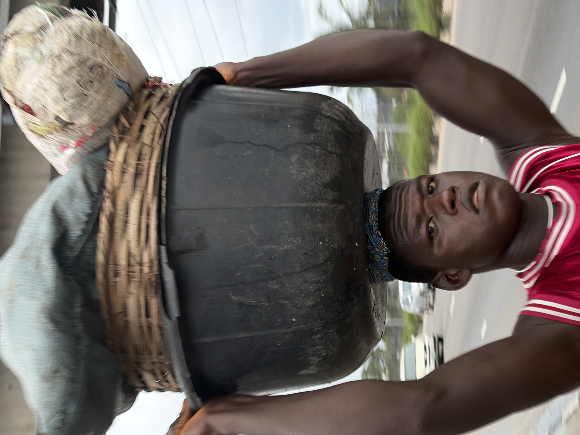 A person, viewed from the chest up, is walking down a road, carrying a large black tub filled with goods on their head. The tub is stabilized by a woven basket and a patterned cloth. The person is wearing a red vest with white stripes. In the background, there are cars and trees, suggesting an outdoor environment. The person's expression is neutral.