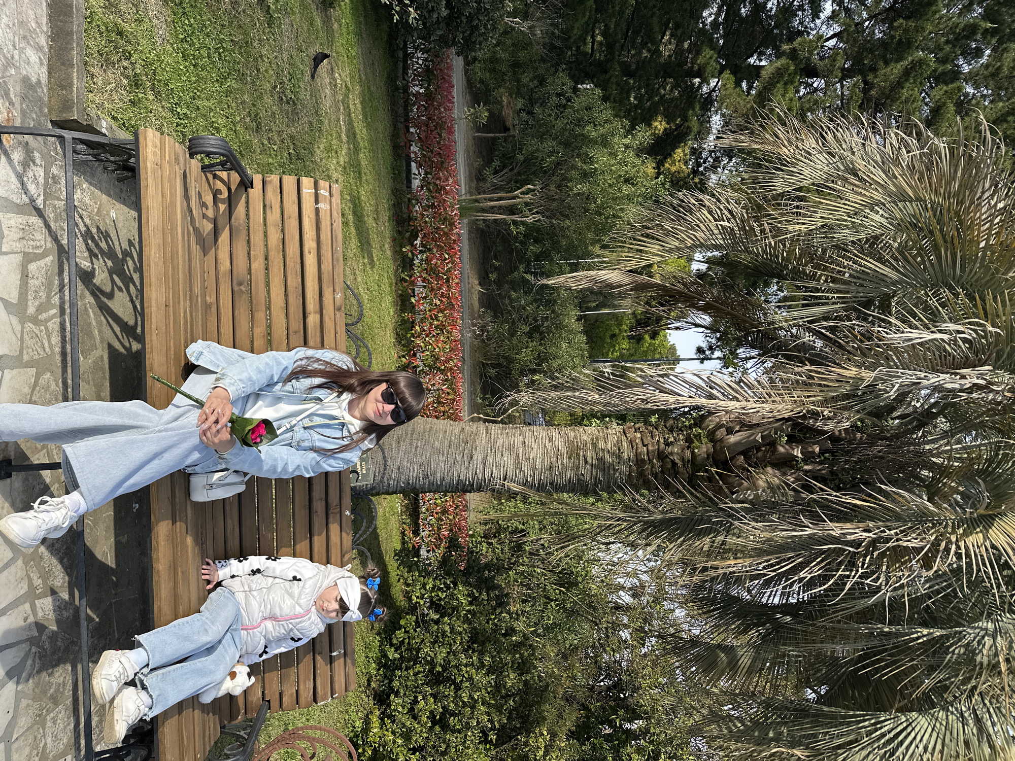 An outdoor scene captured in bright daylight, showing two individuals, an adult woman and a young child, relaxing on a wooden park bench. The image is oriented vertically, with the subjects appearing to be lying on their backs relative to the frame.
The adult woman is seated on the bench, wearing a light blue denim jacket, white hoodie, light-colored wide-leg jeans, and white sneakers. She has long dark hair and wears sunglasses. In her hands, she holds a small bouquet of pink flowers. A small white shoulder bag rests next to her.
Beside her, a young child, likely a girl, is lying down on the bench. She wears a white puffy jacket with black details, light-colored jeans, and white sneakers. Her head is adorned with a white headband and blue hair bows. A small white stuffed animal lies next to her.
The setting appears to be a park or botanical garden, with lush green grass and various trees and bushes. Prominent in the frame are several large palm trees with rough trunks and fronds that show some signs of dryness. A line of bushes with both green and reddish foliage runs in the background. To the left of the bench, there's a paved area made of stone slabs. The bright sunlight suggests clear weather.
On the backrest of the bench, behind the woman's head, a small plaque or sign is visible, with the word "СОЧИ" (Sochi) clearly legible in Cyrillic, consistent with the provided location context of Sochi, Russia. The overall atmosphere is one of leisure and relaxation.