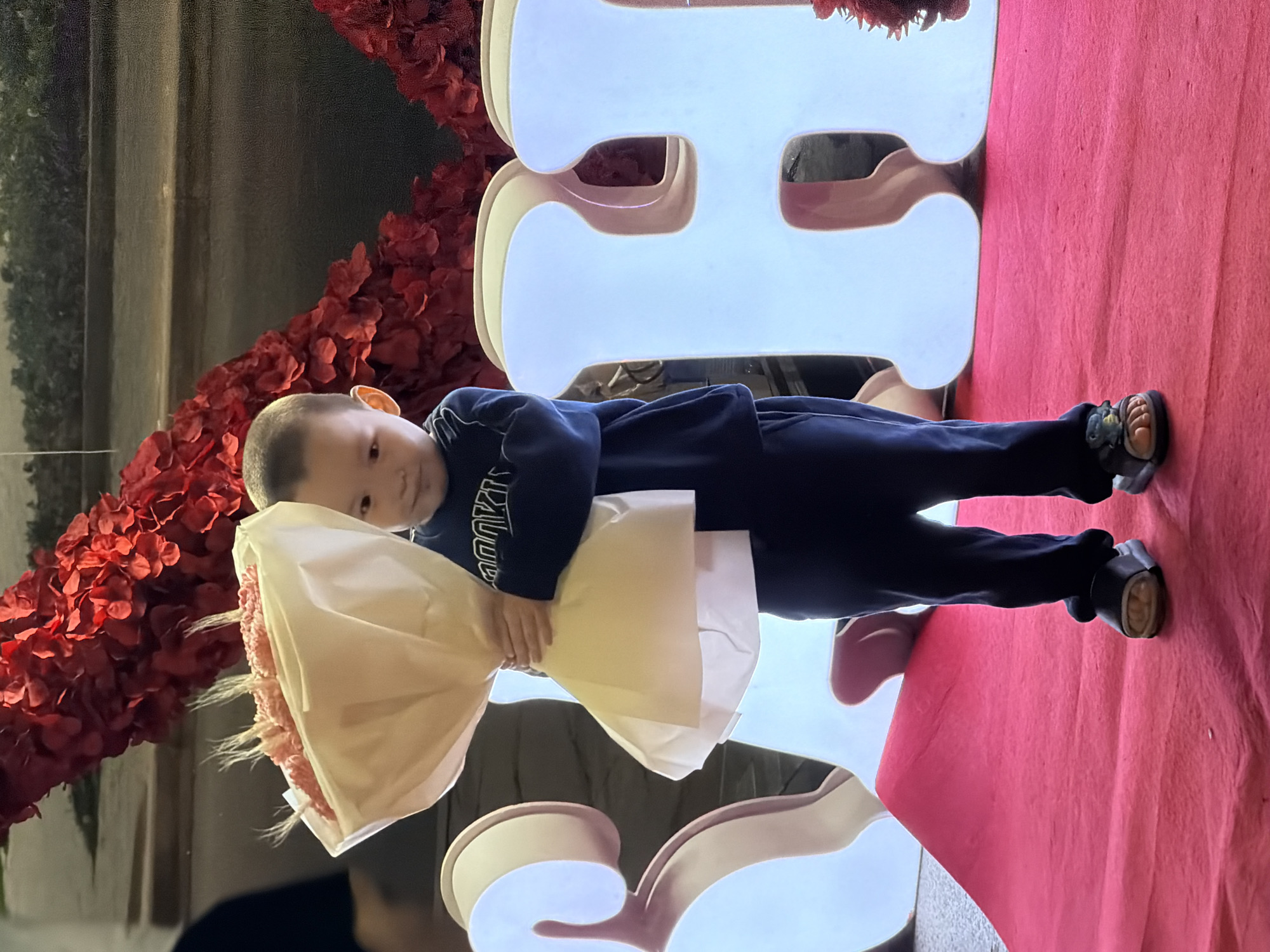 A young boy stands on a red carpet, holding a bouquet wrapped in light-colored paper. He is wearing a dark blue sweatshirt and pants, and dark sandals. Behind him are large, illuminated white letters. Red floral decorations are visible in the background. The setting appears to be an indoor event.