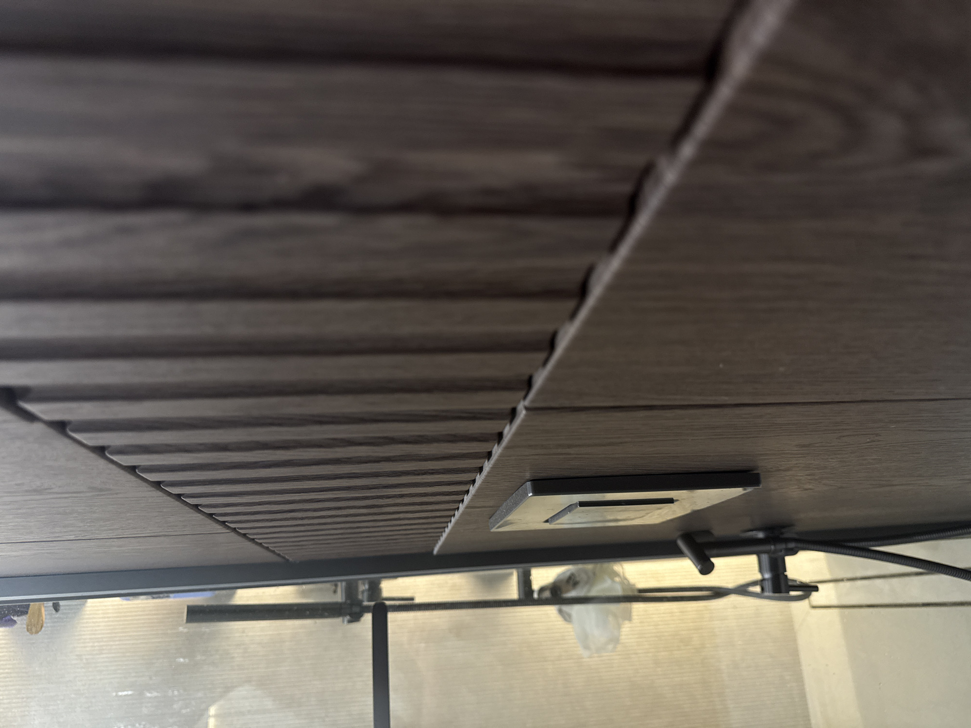 The image provides a low-angle view looking up at the underside of an overhead structure, likely an outdoor deck, patio cover, or a ceiling in a covered space. The primary subjects are several interlocking rectangular panels with a dark greyish-brown wood-grain texture. Some panels are smooth, while others feature distinct horizontal ribbing.
Below these panels, a dark metal frame provides support. Attached to one of the flat panels is a small, rectangular metallic device, possibly an integrated light fixture or a sensor. Further to the right, a black cable or hose extends from a cylindrical black fixture, which also has a small black rod protruding from it.
The background, seen through or beyond the metal frame, is blurred but appears bright, suggesting daytime. It shows indistinct shapes and a translucent white object, possibly a plastic bag or cover, hanging or positioned in the distance. The overall impression is a modern, functional architectural detail. No people are visible, and no specific activity is taking place. There is no discernible text in the image. The setting is an architectural environment, with this particular scene located in Chaikovsky, Russia.