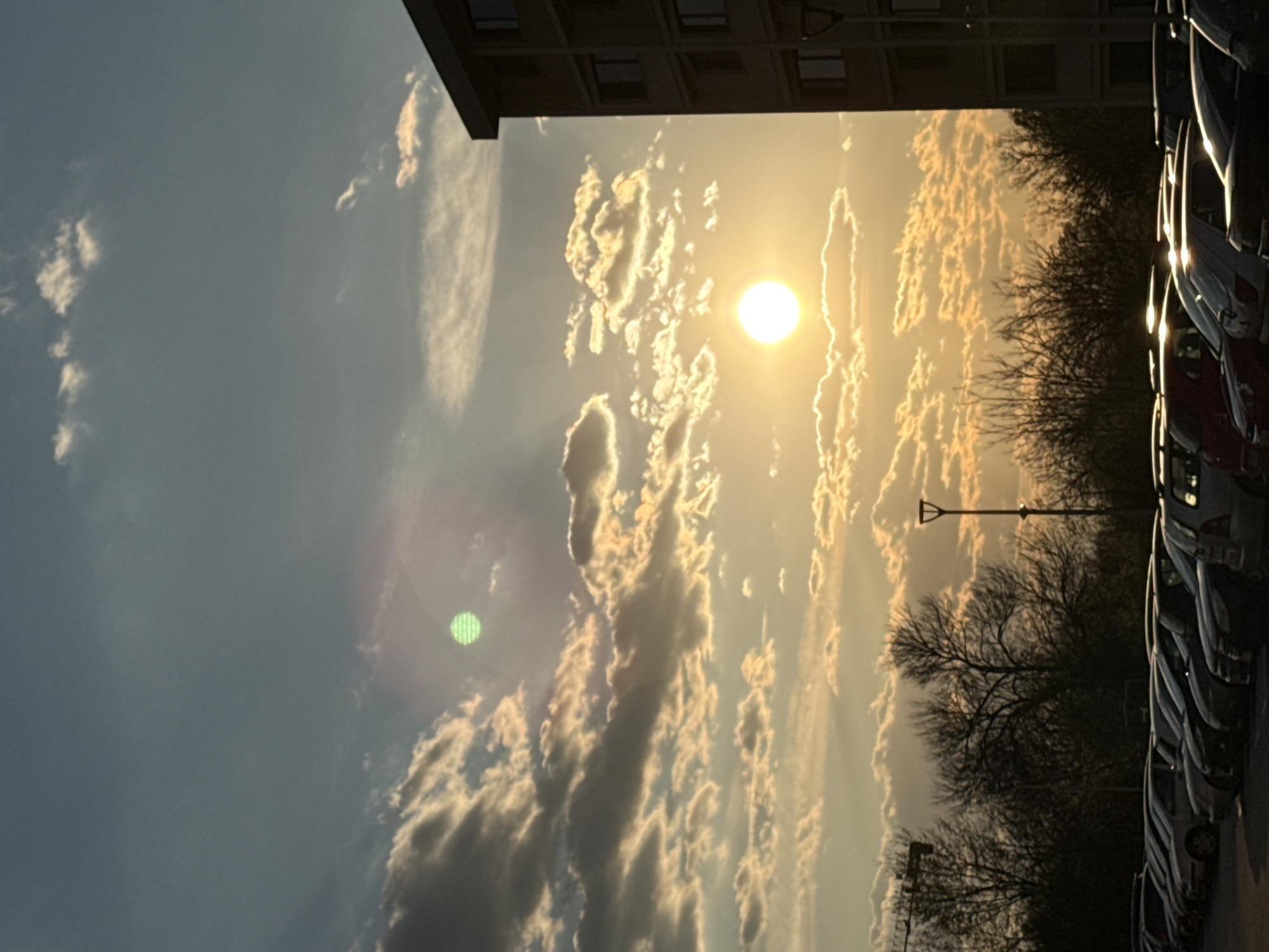 The image shows a sky at dusk with clouds illuminated by the setting sun. A building is visible at the top. Trees line the right side of the image, along with a row of parked cars. A lamppost stands among the trees. A green lens flare is visible in the sky. The location is in a city; the specific city is Unknown, Unknown.