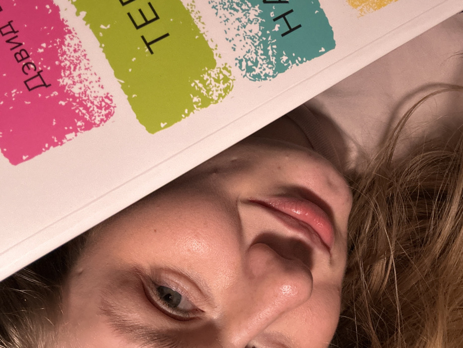 A person lies on their side, a book partially covering their forehead. The book has colorful paint strokes with Cyrillic text. The person has fair skin, blonde hair, and a single eye visible, looking toward the viewer. Their lips are slightly parted. The background is a soft, light color, possibly a shirt or bedding. The lighting is soft, with some shadows highlighting the facial features.