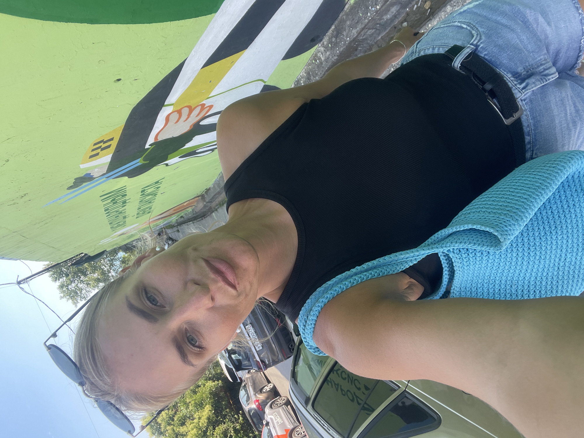 The image shows a woman posing for a selfie outdoors. She has blonde hair and is wearing a black tank top and denim shorts. She is holding a light blue knitted bag over her shoulder. In the background, there is a wall with a colorful mural, a line of parked cars, and trees. The sky is visible, suggesting it is daytime. The overall scene appears casual and summery. Some text is visible on the mural and on the side of a car, but it is not clearly legible.