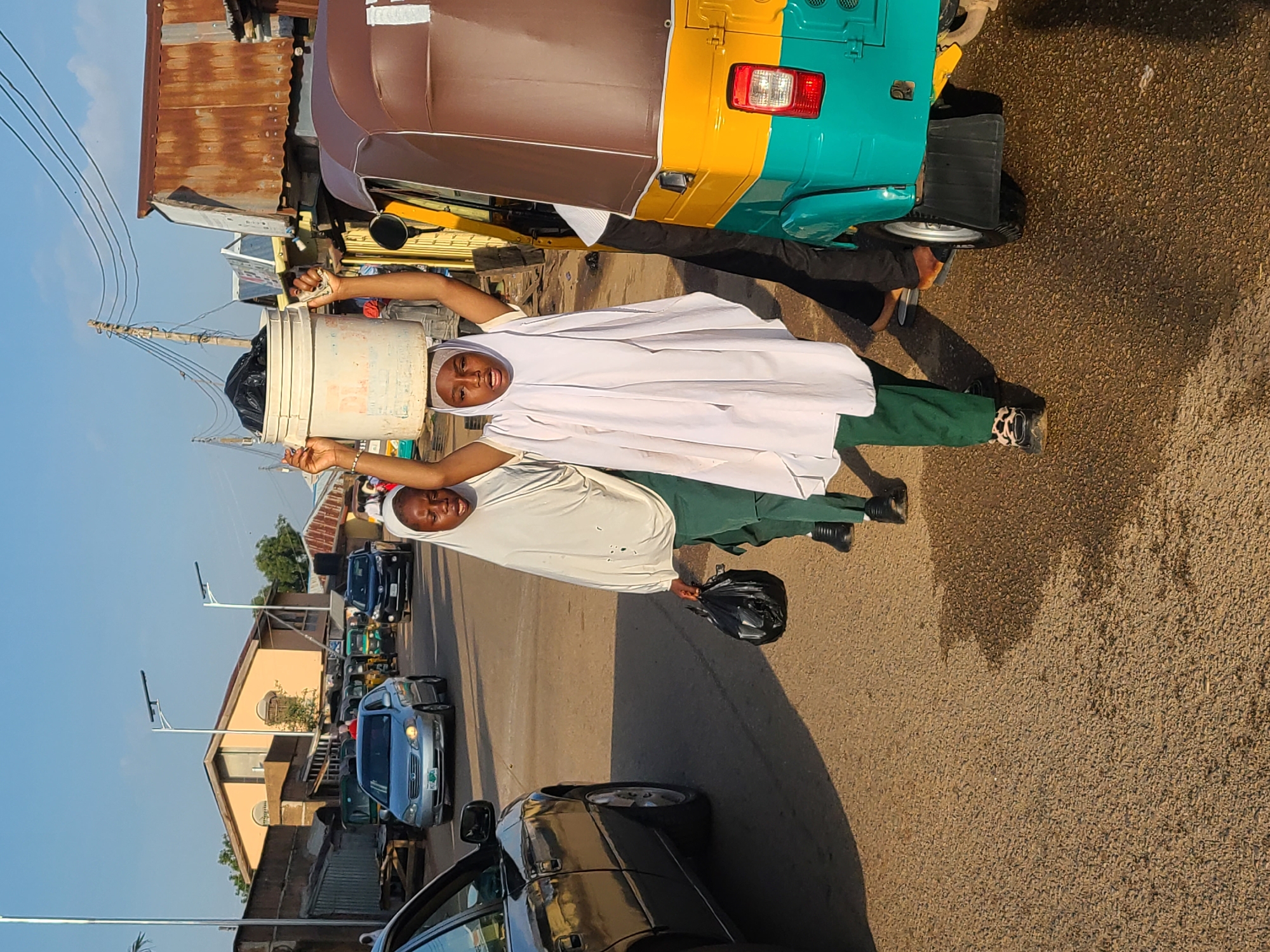 In Jos, Nigeria, on a sunlit street, two young girls are walking. The girl in front, wearing a white hijab and green pants, carries a large white bucket filled with black plastic bags on her head. She also holds a black plastic bag in her right hand. The second girl, walking slightly behind and to her left, wears a similar white hijab and green pants and carries a black bag in her left hand. They are on a paved road with vehicles, including a silver car and a colorful auto-rickshaw. Buildings and shops line the street, and overhead, electrical wires are visible against a clear blue sky. The scene depicts a moment of everyday life, possibly related to commerce or daily chores, in an urban environment.