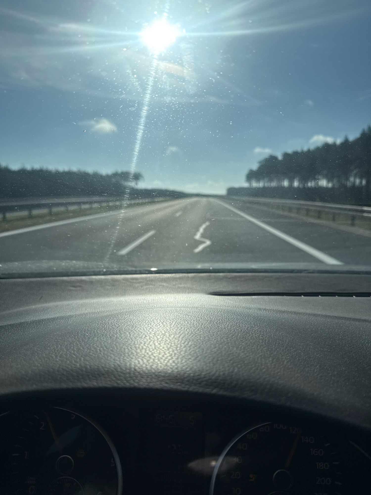 The image shows a view from inside a car traveling on a highway. The sun is shining brightly, causing glare on the windshield and some lens flare. The road ahead is clear, with a solid white line on the right and a broken white line on the left. Trees line the sides of the highway in the distance. The dashboard of the car is visible in the foreground, with a speedometer showing a speed of approximately 120 km/h. There is no visible presence of people or specific landmarks that would indicate the location in Trzebiszewo, Poland. The environment appears to be rural or exurban, characterized by a highway and natural vegetation.