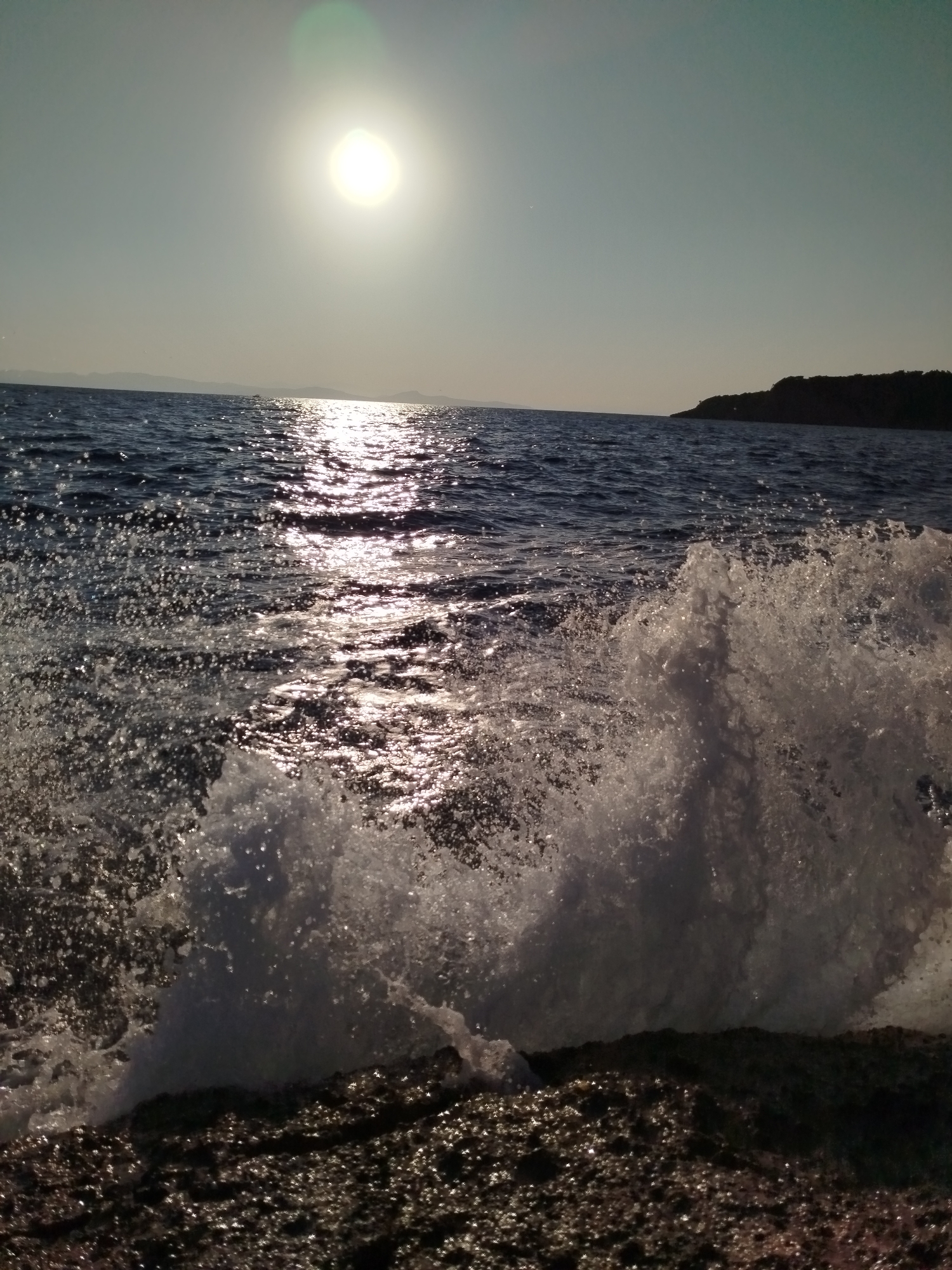 A dramatic, low-angle shot captures a wave crashing against a rocky shore at sunset in Karaburun, Turkey. The sun, a bright, diffused orb near the top left of the frame, casts a shimmering, golden path across the dark blue water. A large wave, captured mid-crest, dominates the foreground, its white spray a stark contrast against the dark rocks and water. The rocky shore is wet and glistening, reflecting the sunlight. In the distance, a silhouetted headland, likely part of the Karaburun peninsula, juts out into the sea under a pale, clear sky. No people are visible in the image.