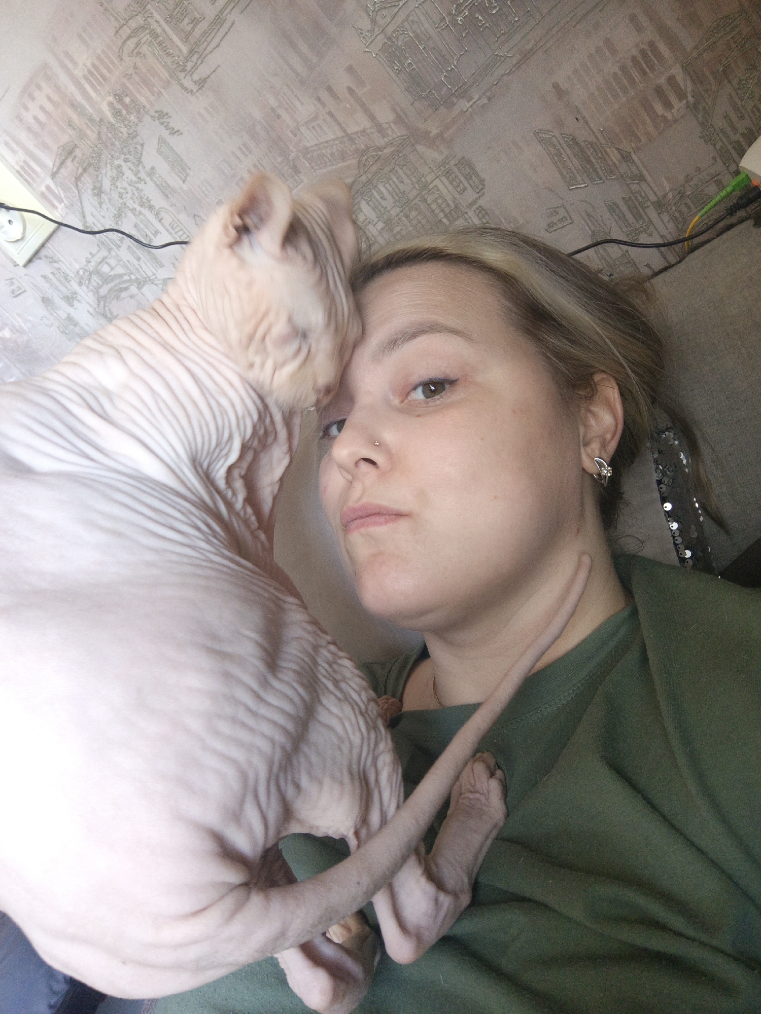 A woman is pictured in a selfie with a Sphynx cat. The woman has light skin, light brown hair with blonde highlights, and wears a small nose ring in her left nostril and silver hoop earrings. She is wearing an olive green t-shirt. The cat is hairless, with wrinkled skin, and has its head resting on the woman’s head. The cat’s tail extends along the woman's neck and down her chest. The background is a textured wallpaper. A white electrical outlet with wiring is visible in the upper left corner, and more wires are seen towards the upper right. The location may be in Марковский, Russia, though this cannot be confirmed from the image itself.