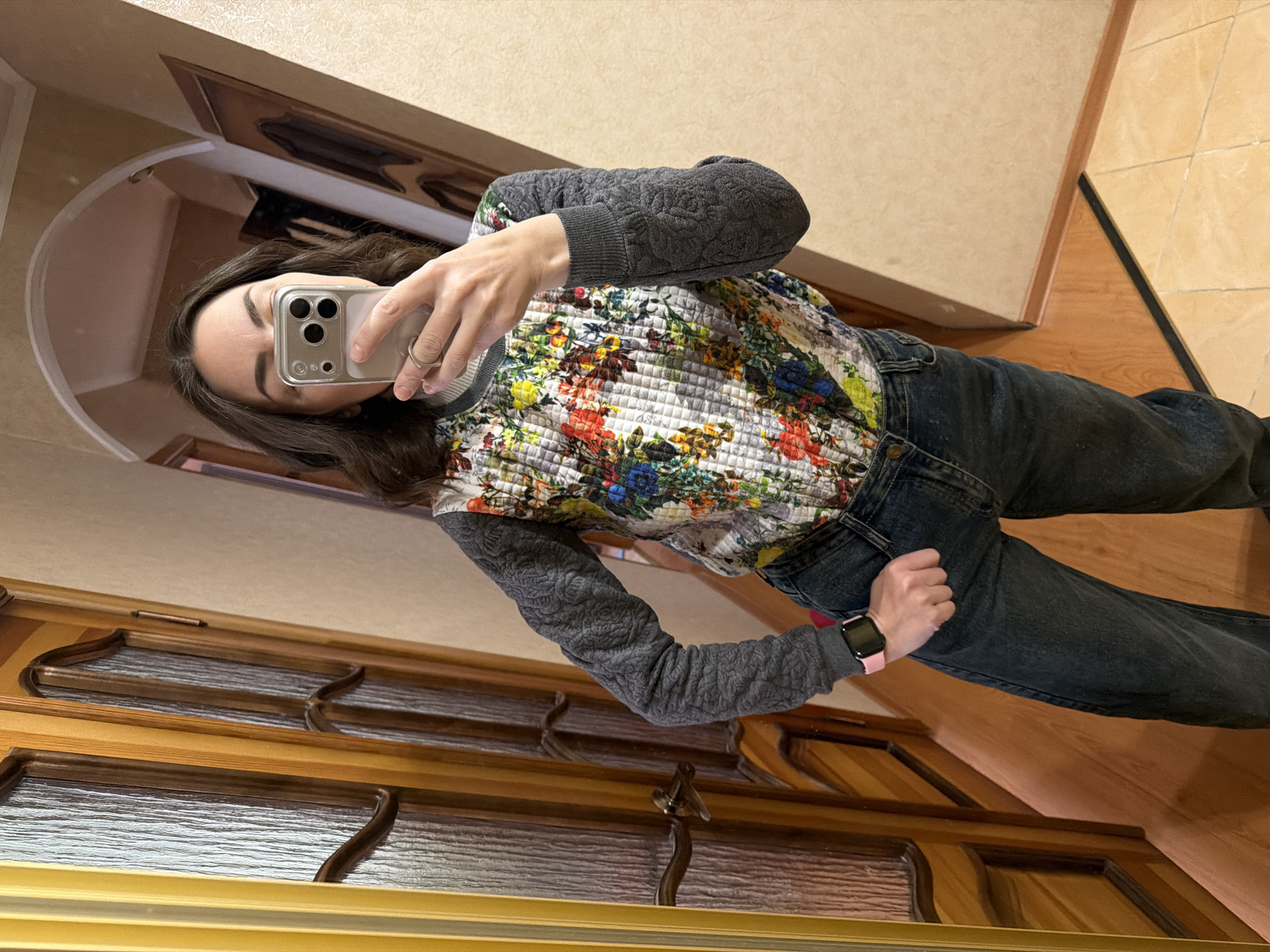 A person is taking a mirror selfie in an indoor setting. The individual is wearing a floral patterned top with gray quilted sleeves and dark jeans. They are holding a smartphone in front of their face, obscuring it from view. A watch with a pink band is visible on their wrist. The background includes a wooden door with ornate glass panels, a light-colored wall, and a section of tiled flooring. The exact location within the city of Unknown, Unknown is not discernible from the image.