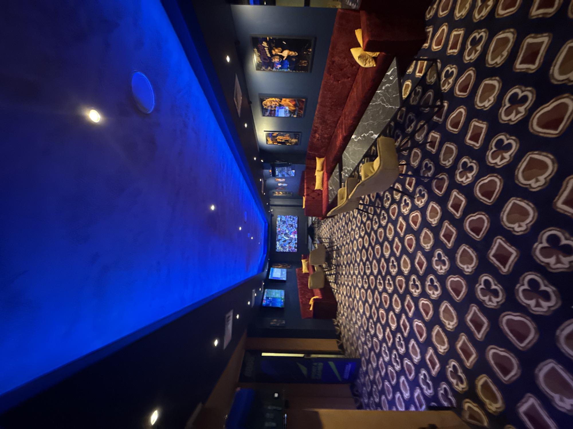 The image shows the interior of a long room, possibly a bar or lounge, in Burgas, Bulgaria. The left side of the room features a striking blue illuminated ceiling that extends along the length of the space. Adjacent to this is a dark wall adorned with framed pictures.
On the right side, there is a long red upholstered bench with yellow cushions. In front of the bench are black marble tables with tan-colored chairs.
The floor is covered with patterned carpeting with symbols of playing cards: spades, hearts, diamonds, and clubs. Toward the end of the room are multiple television screens displaying various images. The overall lighting appears to be dim with accents of blue and warm tones from the interior lighting.