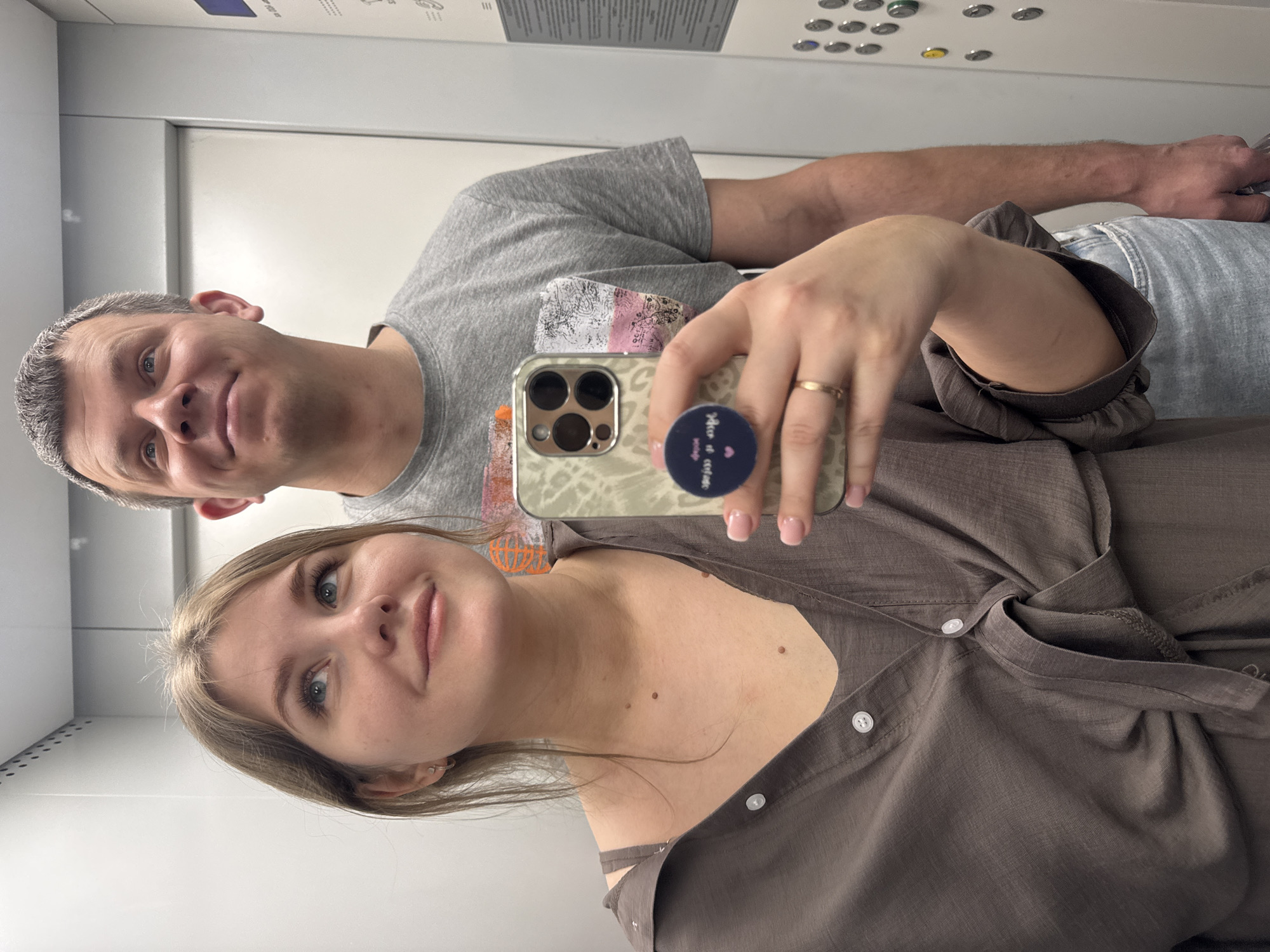 A couple takes a selfie in an elevator. The woman is holding the phone, a gold-colored iPhone with a patterned case and a blue pop socket. She is wearing a brown button-down shirt. The man is wearing a grey t-shirt and denim jeans. The elevator interior is plain with a button panel visible in the background. The location appears to be Chaikovsky, Russia, based on context.