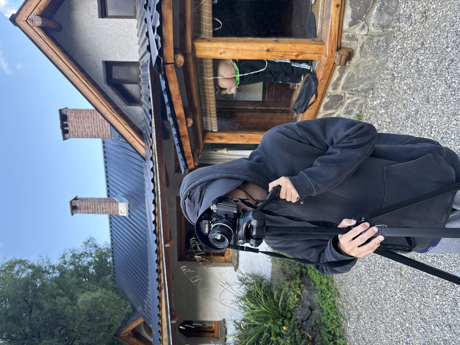 A person wearing a dark gray hooded sweatshirt is holding a vintage Nikon camera on a tripod, aiming towards a building. The building has a wooden frame and a stone facade. A person is visible in the window, wearing black pants with a green stripe. The background includes a blue sky and green foliage. The camera is the focal point of the image, with the building serving as a secondary element.