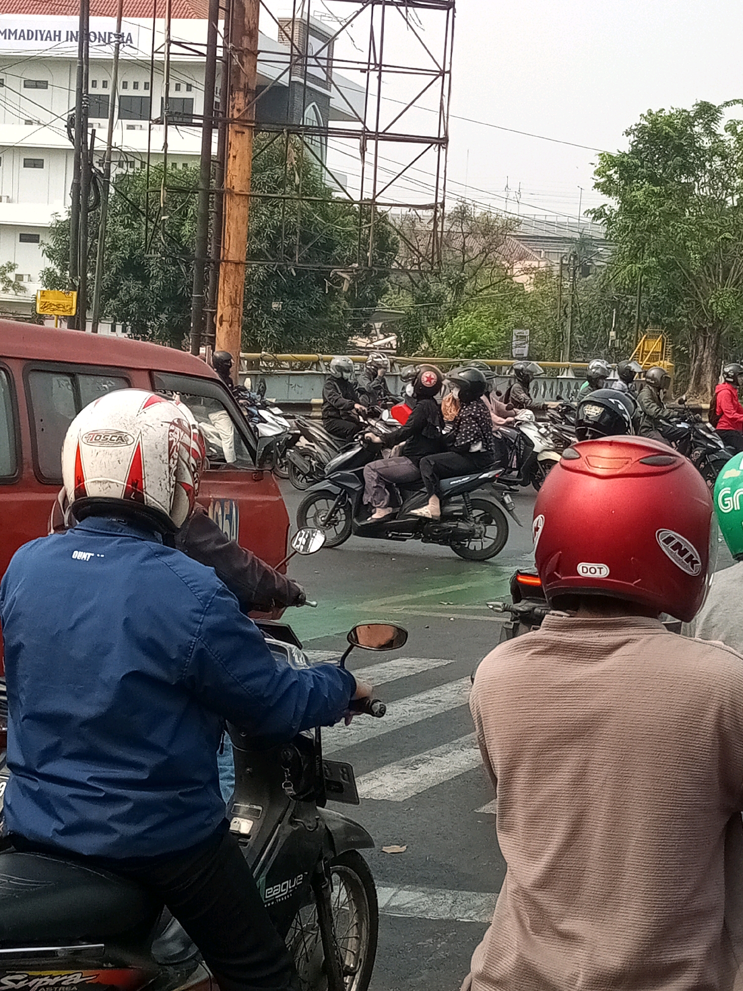 In Bekasi, Indonesia, a traffic jam is depicted with a multitude of motorcycles and a red van. The foreground features two individuals on motorcycles, both wearing helmets. The person on the left wears a blue jacket and a white helmet with red and white markings. The person on the right wears a brown shirt and a red helmet. Several other motorcyclists, many in helmets, are visible in the mid-ground, queuing behind a red van. The environment includes trees, a building with the text "MMADIYAH INDONESIA" visible, and a large metal structure, possibly an overhead sign or scaffolding. The scene is set on an asphalt road with white crosswalk markings.