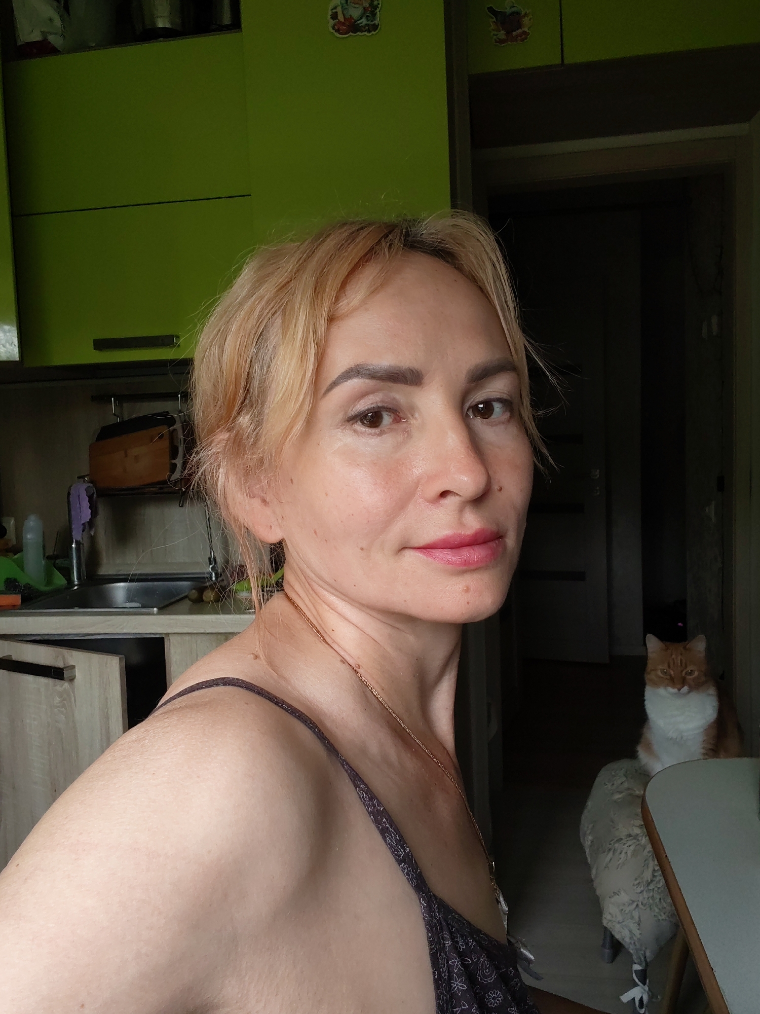 A medium shot captures a woman with blonde, medium-length hair, wearing a dark-colored, patterned spaghetti strap top, looking towards the camera with a neutral expression. She has fair skin, prominent cheekbones, and light brown eyes. A subtle pink lipstick is applied to her lips. She is positioned in the foreground, with her left shoulder and upper arm visible. In the background, an orange and white cat with green eyes sits on a chair. The environment appears to be a kitchen with green cabinets and a sink area visible to the left. The overall scene is indoors, possibly a residential setting, with a doorway and shadowed interior in the background. The location is the city of Chaikovsky, Russia.