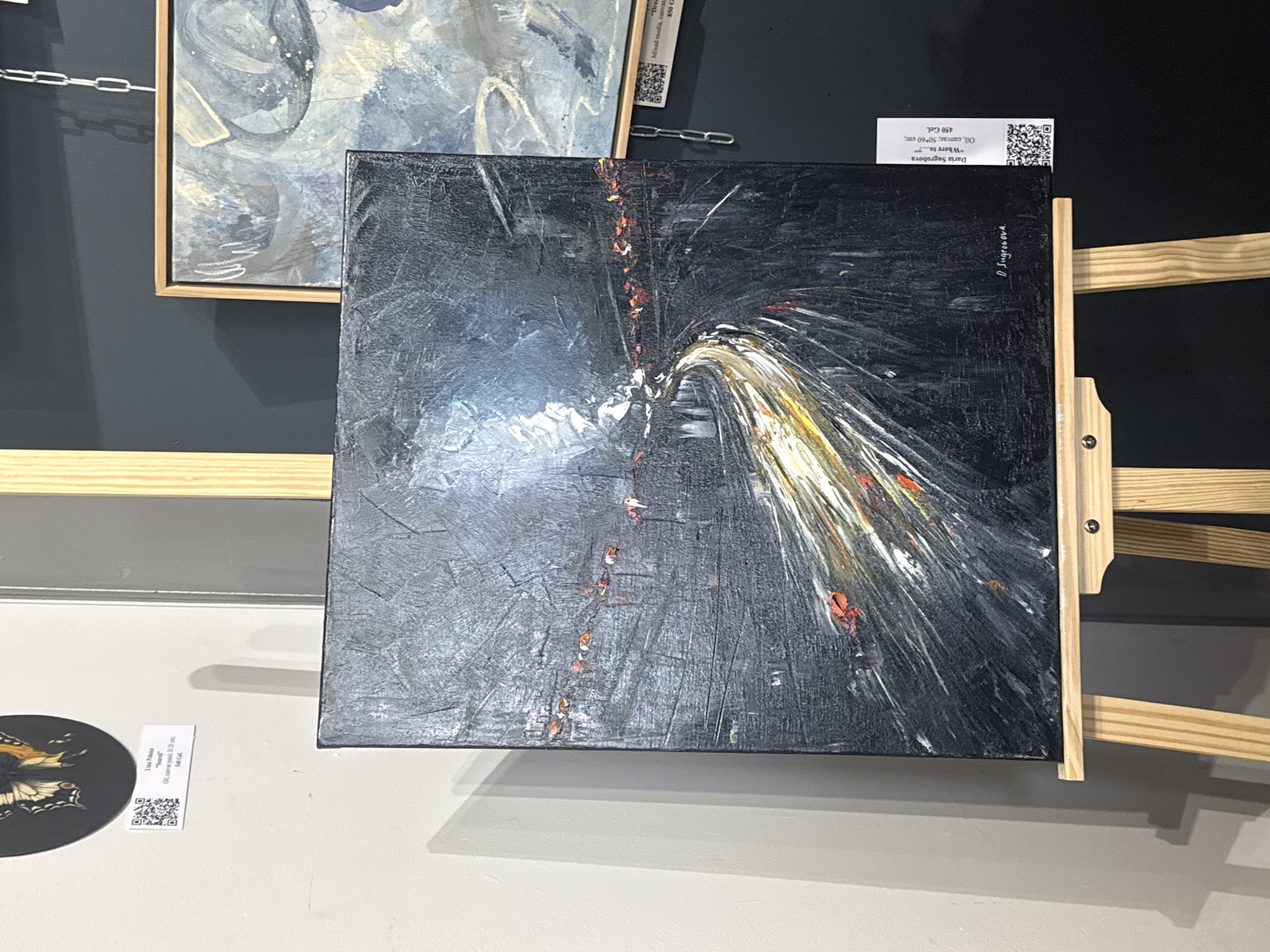The image shows an art exhibition. In the foreground, there's an abstract painting with a dark background and streaks of white, yellow, and red paint converging towards a central point. The painting is displayed on a wooden easel. A label next to it identifies the artist as Daria Sugrobova and the work as "Where to..?", an oil painting on canvas, 50x60 cm, priced at 450 Gel.
Behind the main painting, another framed artwork is partially visible, exhibiting a blue and grey color palette. To the left of the main painting, there's a circular object with a butterfly design and a corresponding label reading "Lina Polans, 'Secret', Oil, canvas print 17*20 cm, $40 Gal". Another label above the main painting mentions "Dre..." and "Mixed media, canvas, 850 G"
The exhibition is likely located in Batumi, Georgia, given the presence of the Georgian Lari (Gel) as the currency on the price tags.