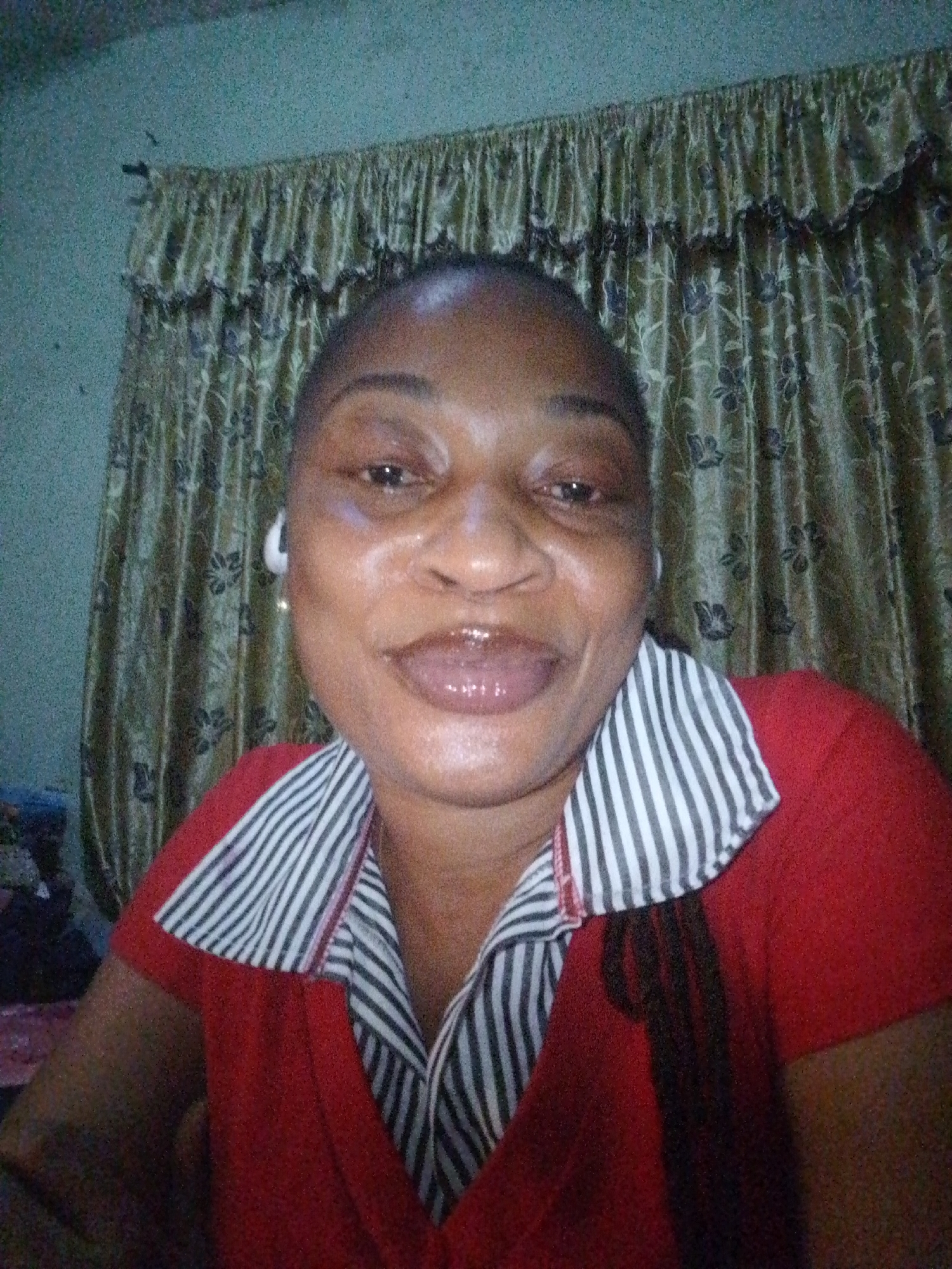 A woman is captured in a close-up, head-and-shoulders selfie taken indoors. She is facing the camera directly with a slight smile and her lips are glossy. She has a light complexion and her head is bald. She is wearing a red shirt with a collar that has black and white stripes. Her left ear is visible and appears to have a white earbud. In the background, there are patterned curtains. The lighting is dim, and the image has a grainy quality. The location is unknown.