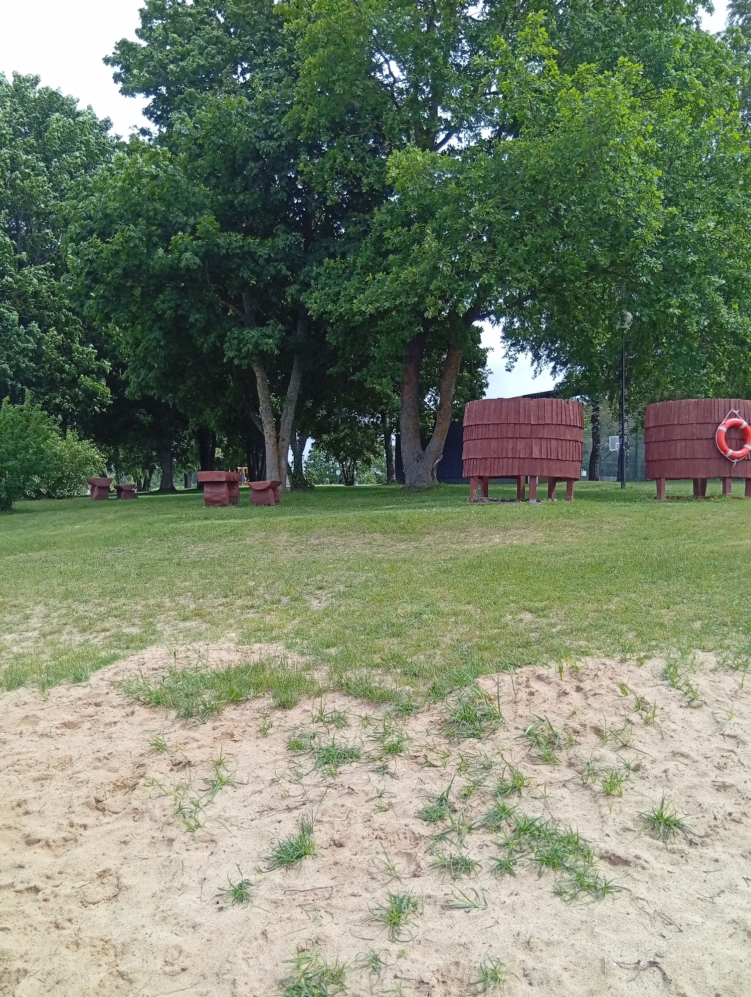 The image depicts an outdoor scene with a sandy foreground and a grassy area beyond. In the grassy area, there are several wooden structures that appear to be benches or tables with attached seating. Further in the background, two large, cylindrical wooden structures are visible, one of which has an orange life preserver attached to it. These structures are elevated on wooden legs and are situated amidst lush green trees. The environment suggests a park or recreational area, potentially near a body of water given the presence of the life preserver. There are no people or animals visible in the frame. The overall impression is one of a calm, natural setting.