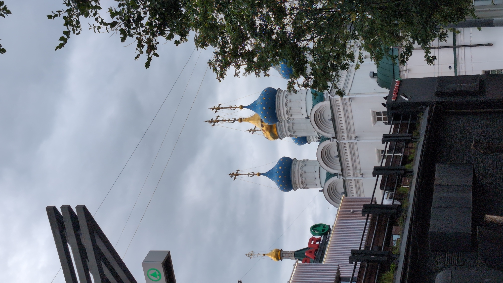 The image shows a street view in Nizhny Novgorod, Russia, featuring a church with blue and gold domes. In the foreground, there is a modern building with a SPAR logo and a black facade. A tree is visible on the right side of the frame, and power lines crisscross the sky. The sky is overcast with clouds.
