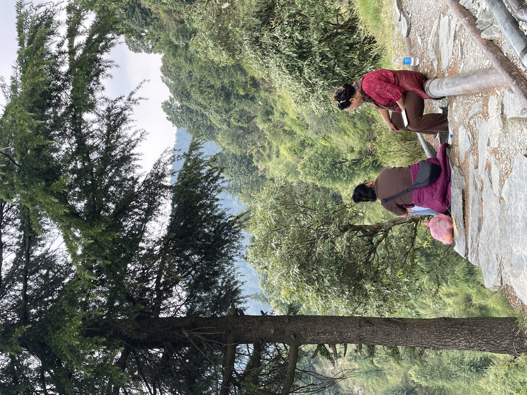 The image captures a scene in Bhagsu, India. Two women are seen on a rocky path. One woman is sitting on a pipe, with a water bottle nearby. The second woman stands nearby. A tree occupies the left side of the frame. The background features lush green vegetation.