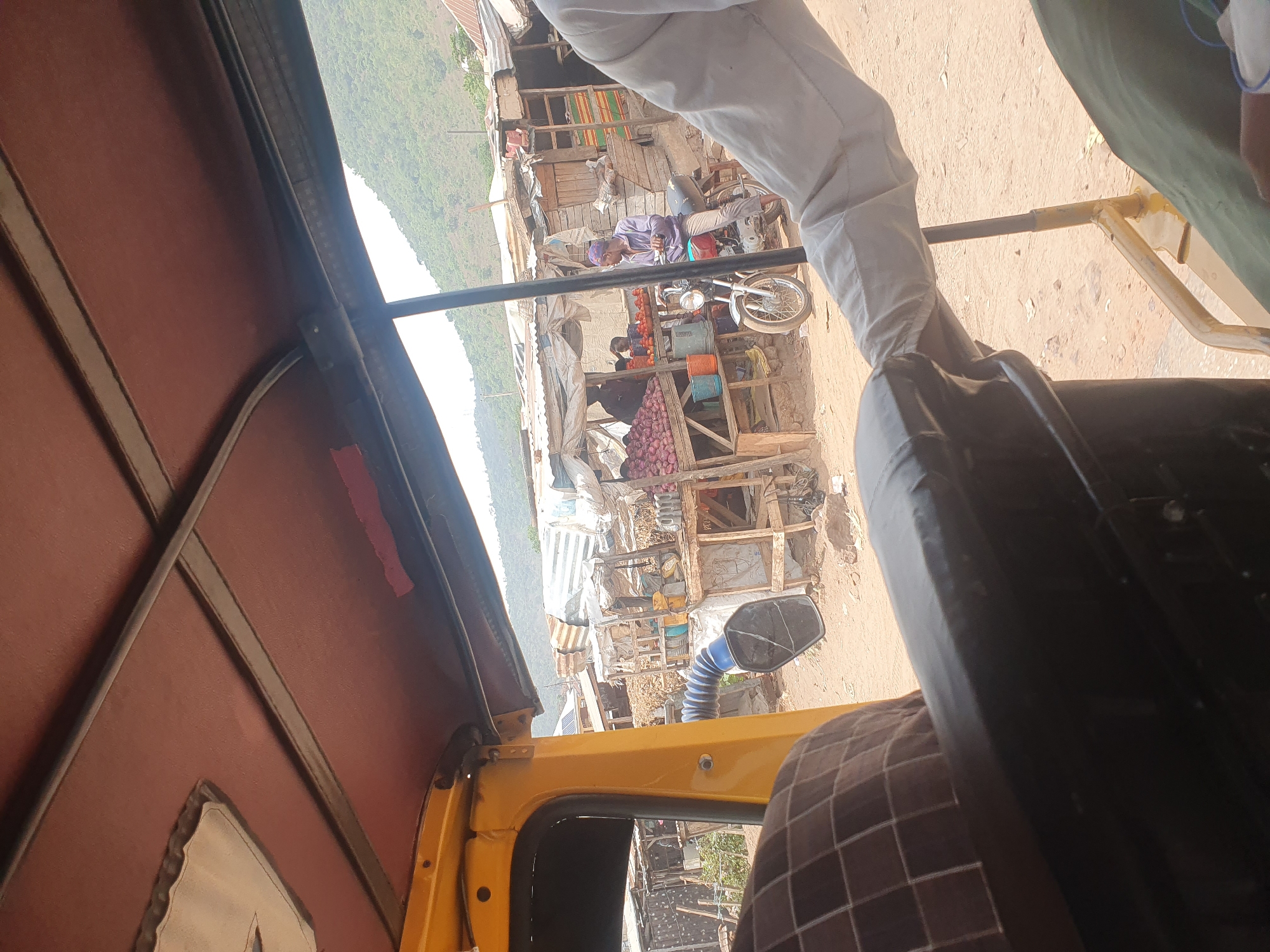 A street scene in Lokoja, Nigeria, is viewed from inside a yellow auto rickshaw. The rickshaw's interior features a red fabric roof. Outside, makeshift stalls and corrugated iron roofs dominate the landscape. A man in purple clothing is seen near a motorbike parked outside one of the stalls. Fruits and vegetables are displayed for sale. The ground is sandy. In the background, a tree covered mountain slope is visible.