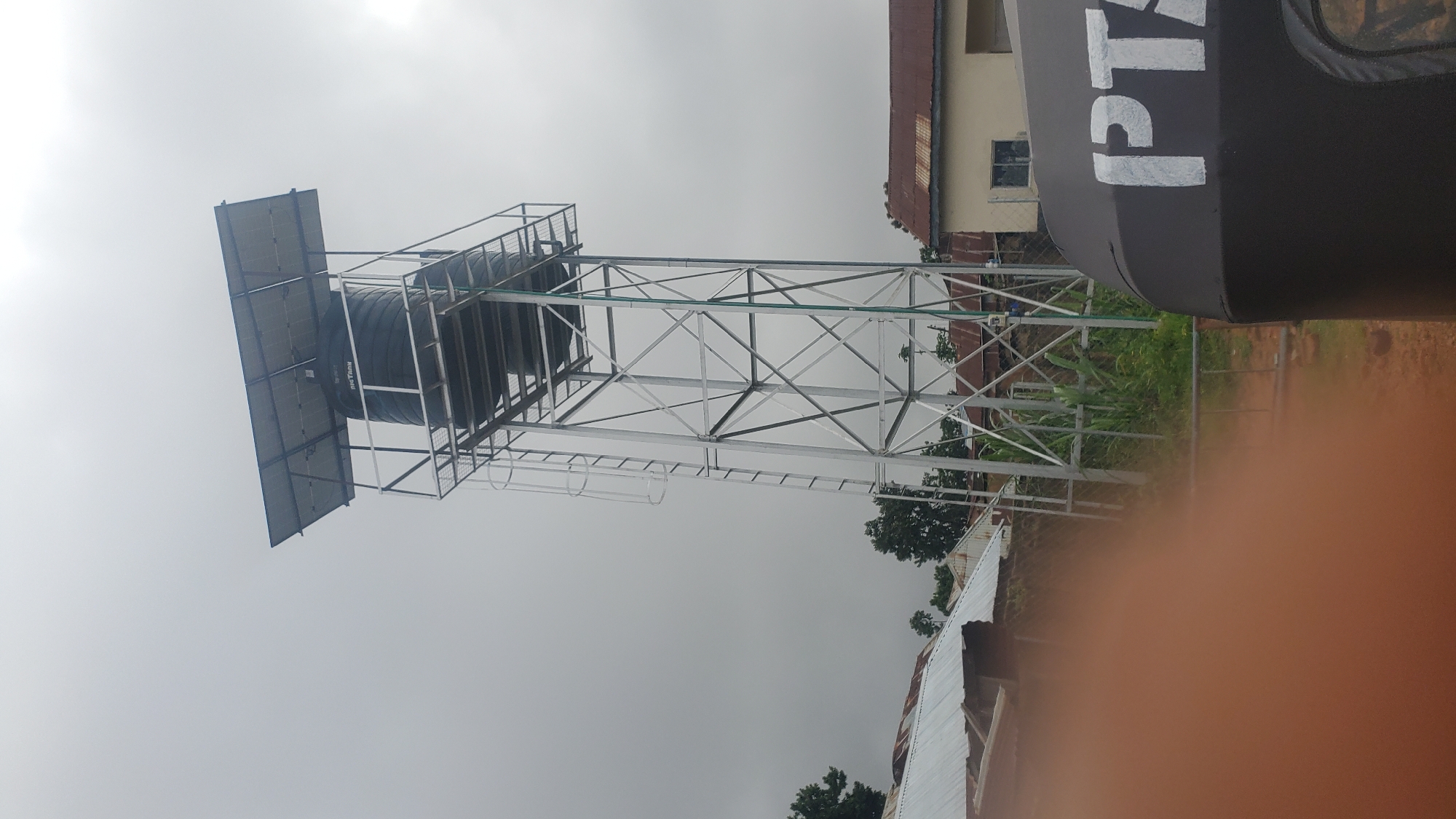 A tall, silver metal water tower with a black water tank and solar panels on top stands in a residential area. In the foreground, the rear of a dark-colored three-wheeled vehicle with the white letters "PT" is visible. Several low-lying buildings with corrugated metal roofs are present behind the water tower. The sky is overcast. The image appears to have been taken in the city of Jos, Nigeria.