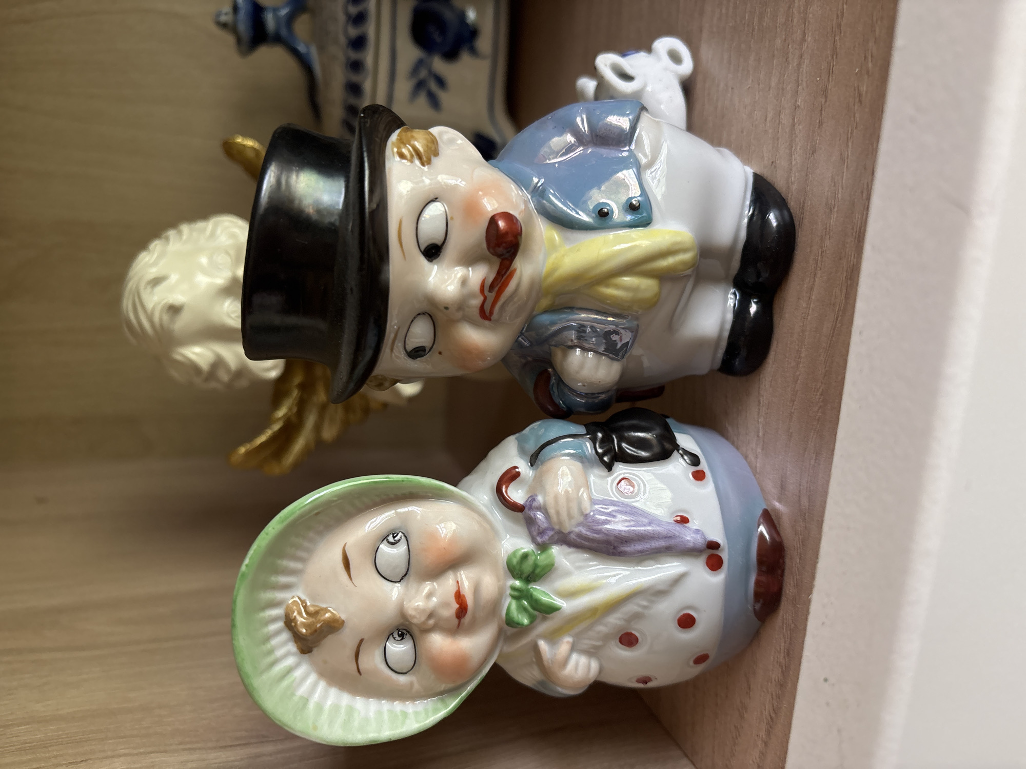 Two ceramic figurines are displayed on a wooden shelf. The figurine on the left depicts a woman wearing a green bonnet, a white coat with red polka dots, and a green bow tie. She holds a closed purple umbrella and a black handbag. The figurine on the right shows a man in a black top hat, blue jacket, and white pants, smoking a pipe. He wears a yellow scarf. In the background, a white ceramic angel with gold wings is partially visible, along with a blue and white patterned decorative box. The scene is indoors, in what appears to be a home setting. The location of Borki, Russia is not visually represented in the image.