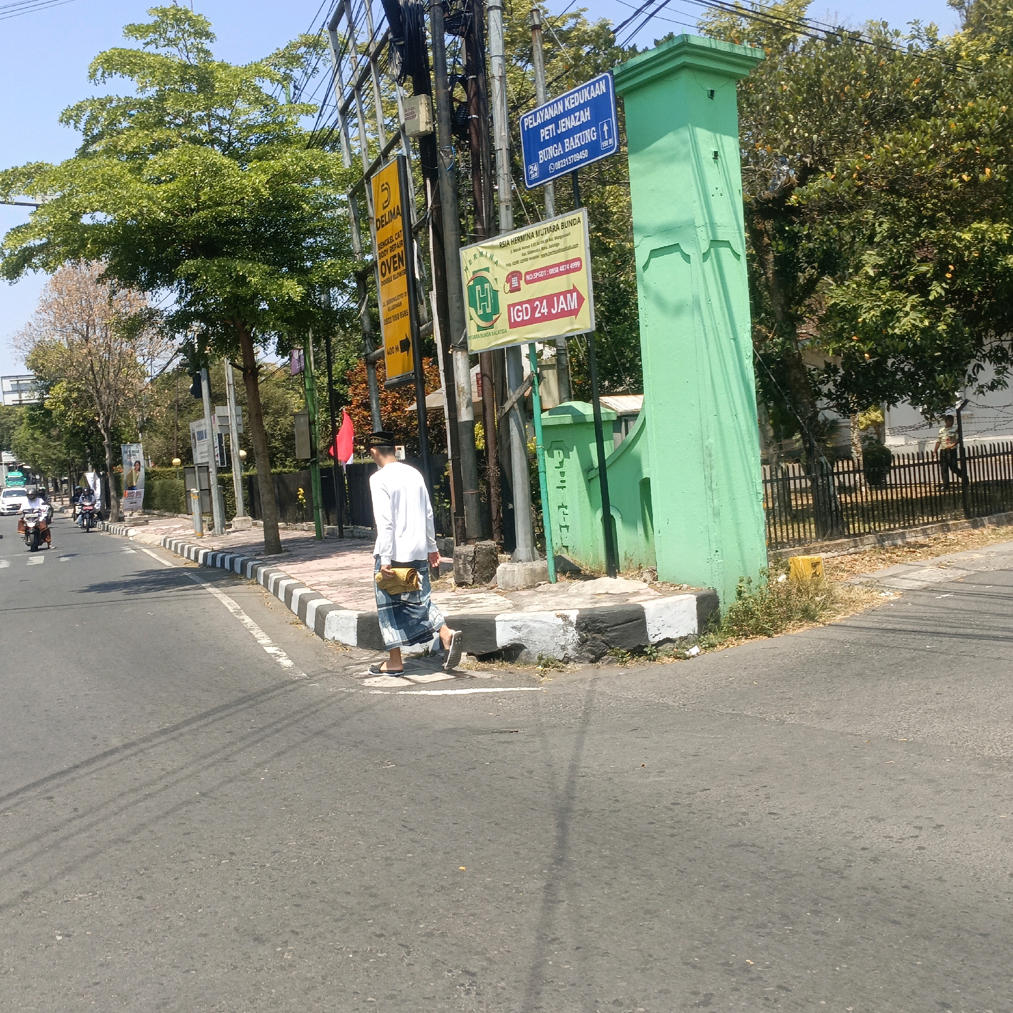 The image shows a street scene in Salatiga, Indonesia. A man wearing a white long-sleeved shirt, a sarong, and a cap is walking on a sidewalk. He is carrying a rolled-up item in his left hand. He is walking away from the camera, towards the right side of the frame. The sidewalk has a black and white striped border. Behind the man, there are several signs on poles. One sign is blue and says "PELAYANAN KEDUKAAN PETI JENAZAH BUNGA BAKUNG" with a phone number and distance. Another sign is yellow and has a "5" logo, with text "DELIMA BENGKEL CAT BODY REPAIR OVEN DOUBLE BLOWER". A third sign is rectangular and yellow with red text that reads "IGD 24 JAM" and a logo with "RSIA HERMINA MUTIARA BUNDA". The green pillar on the right side of the frame is part of a building or structure. Trees line the street, and there are power lines overhead. In the background, there are buildings and traffic, including a few motorcycles and a car. The sky is clear and blue. The road appears to be asphalt.